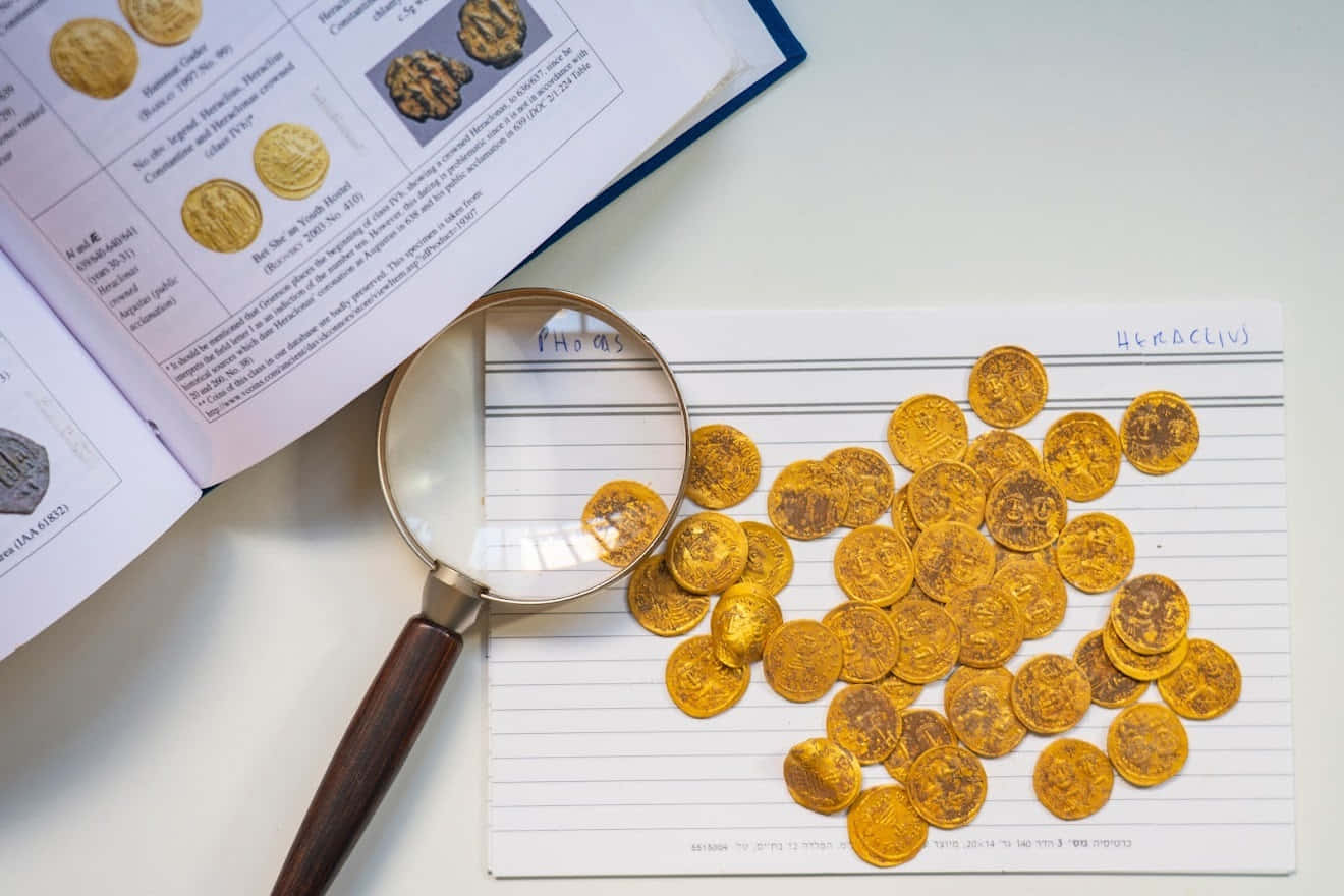 Coin 桌布 壁紙 — 金色古錢幣散落在橫條紙上，旁邊有放大鏡和一本打開的錢幣學書籍。