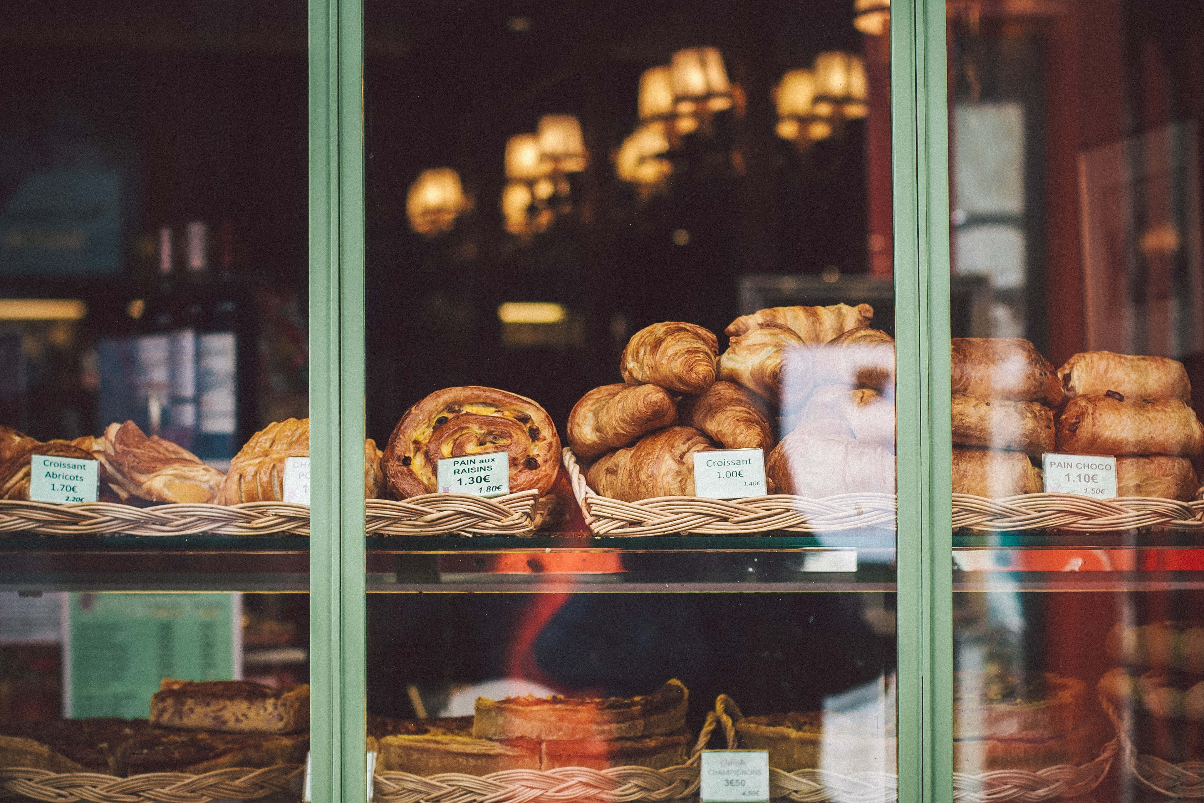 Bakery 桌布 壁紙 — 金色可頌和糕點放在柳條籃子裡，在麵包店櫥窗後，搭配柔和溫暖的燈光。