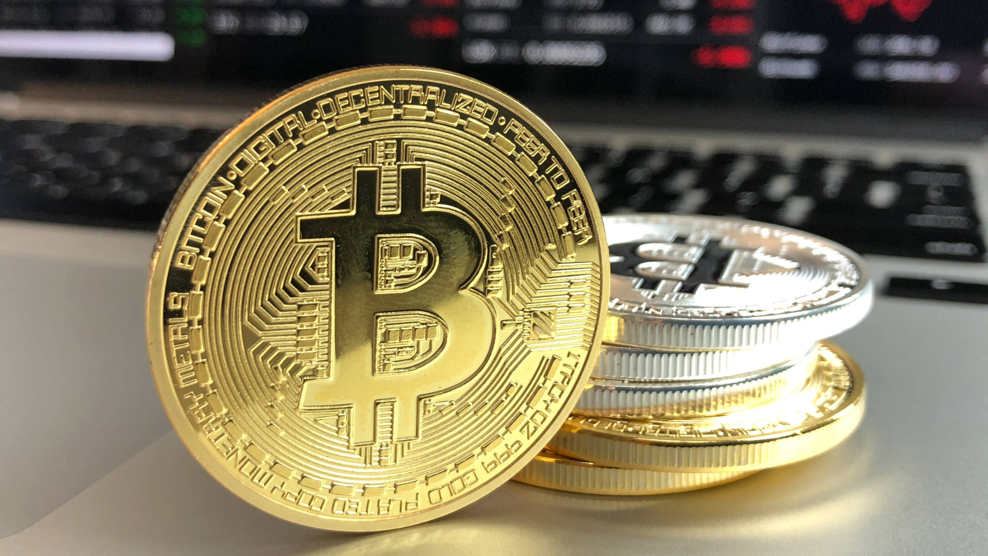 比特幣 桌布 壁紙 — 金色和銀色的比特幣硬幣放在筆記型電腦鍵盤上，背景是發光的股票市場螢幕。