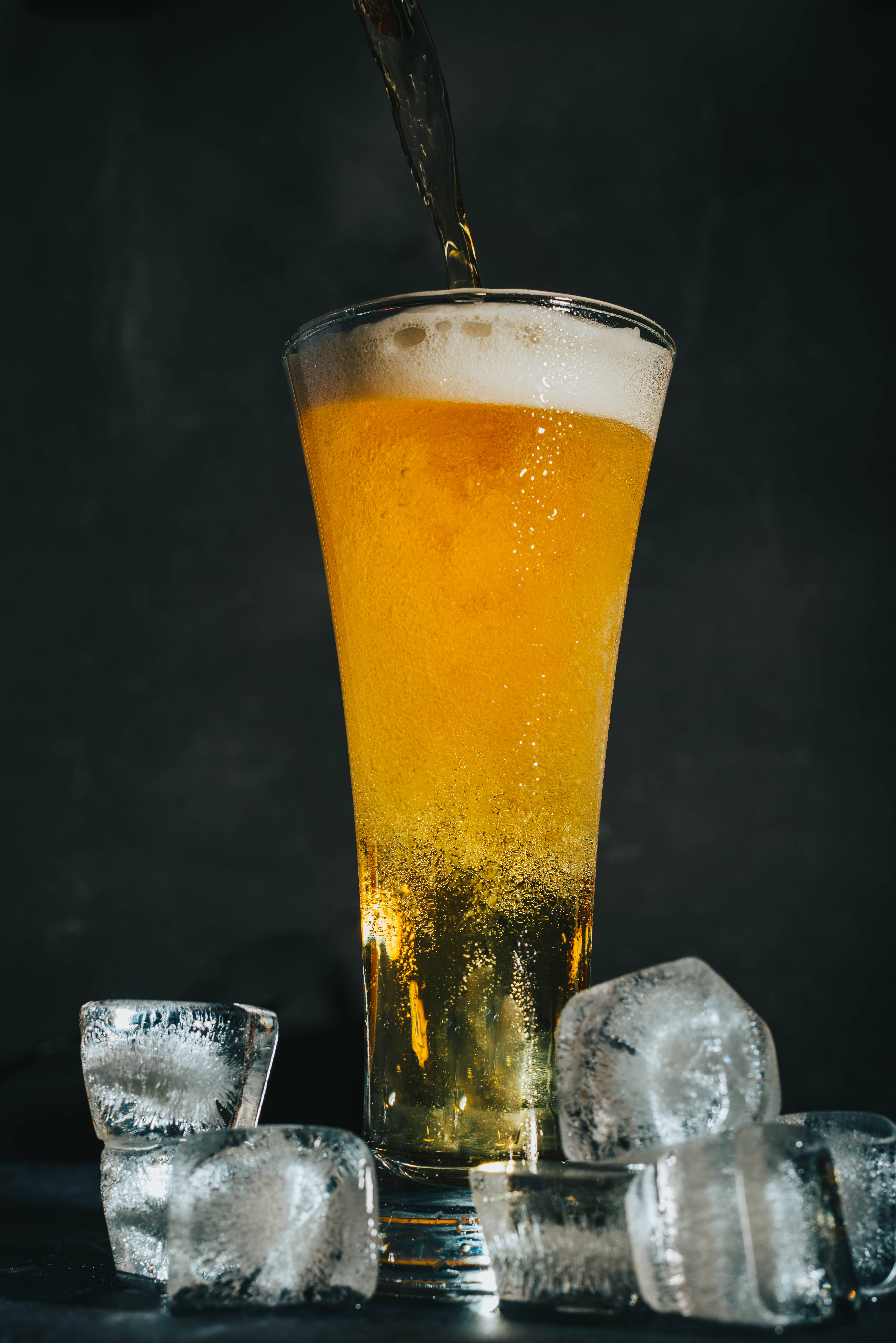 Beer 桌布 壁紙 — 金色啤酒倒入一個高身玻璃杯，周圍有冰塊，背景是深色紋理。