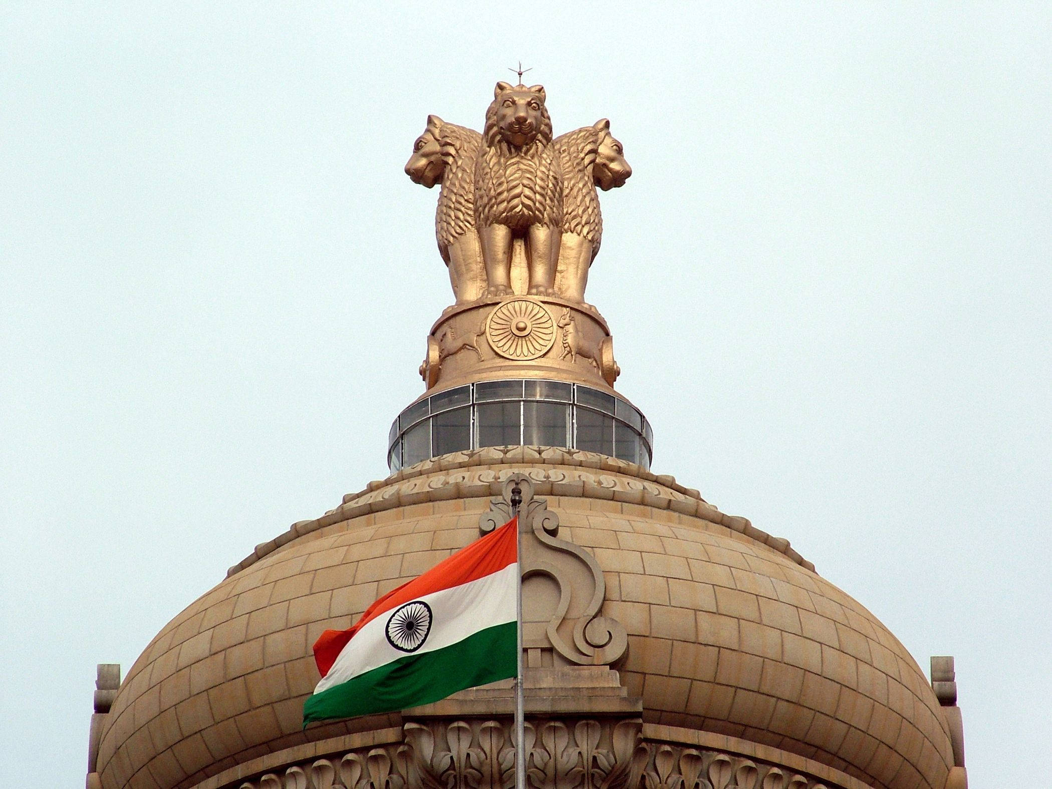 Indian 桌布 壁紙 — 金色四頭獅子雕像矗立在圓頂建築頂端，下方印度國旗在淡色天空下飄揚。