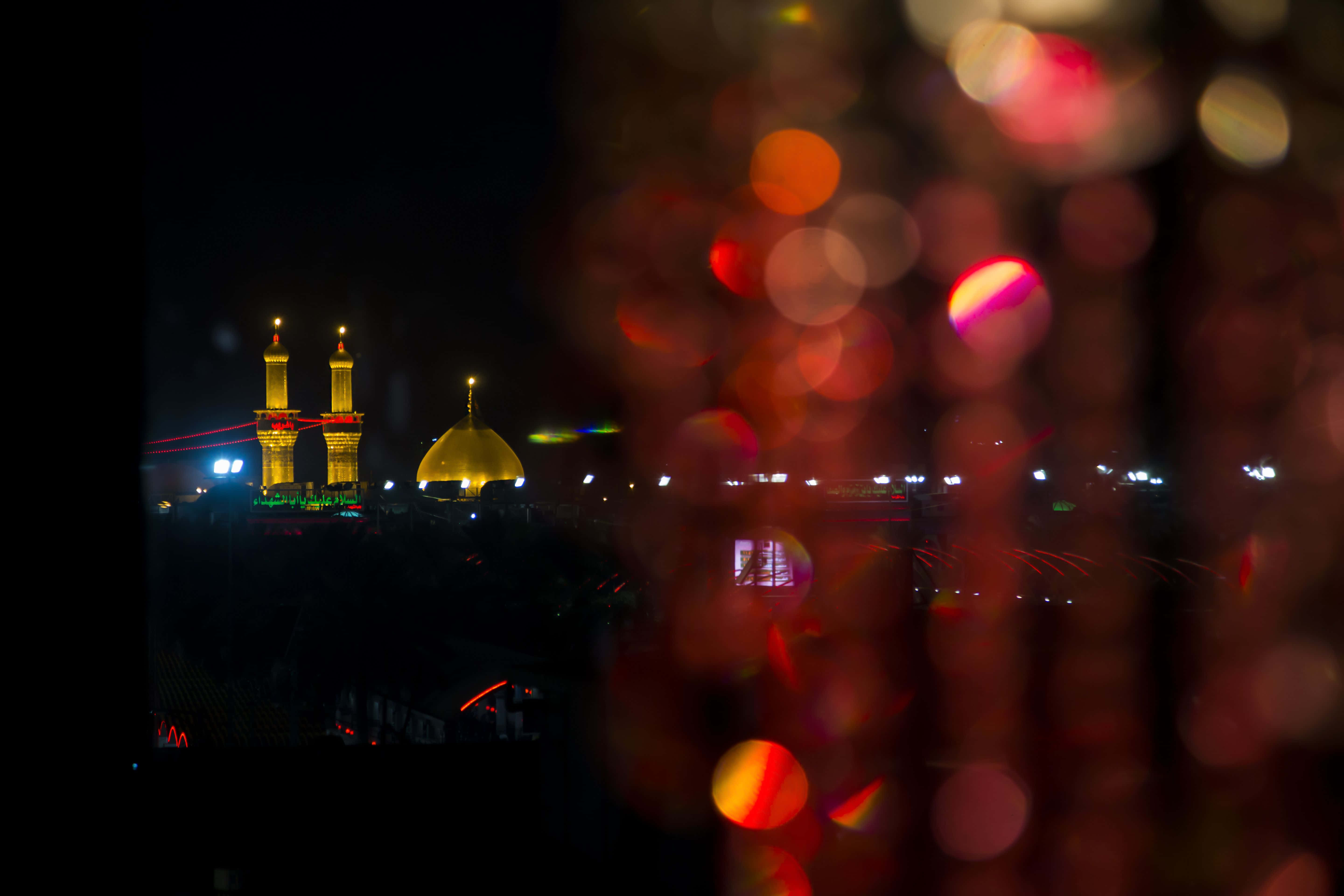 Karbala 桌布 壁紙 — 金色圓頂清真寺和宣禮塔在夜晚透過溫暖的散景燈光前景閃耀。