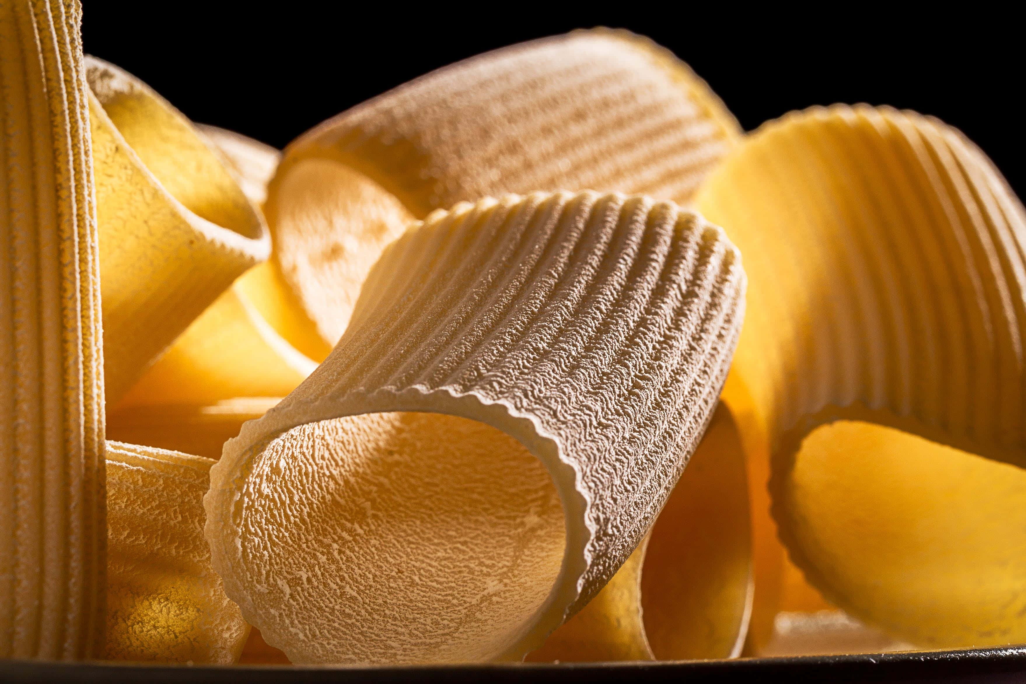 Pasta 桌布 壁紙 — 金色坑紋義大利麵管在溫暖燈光下閃閃發光，襯托著純黑背景。