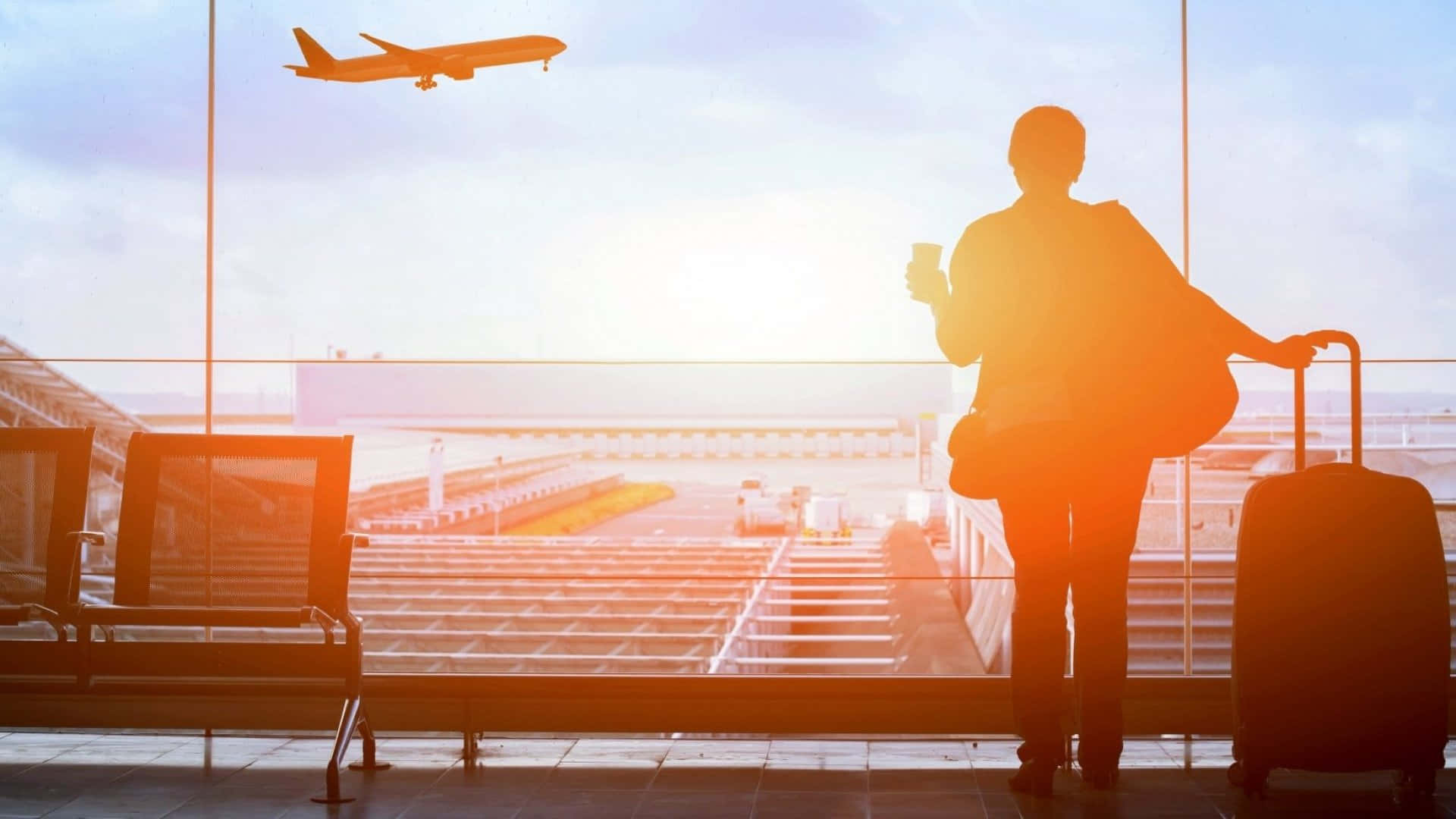 Airport 桌布 壁紙 — 金色夕陽下，一名旅客帶著行李箱透過機場航廈窗戶觀看飛機起飛的剪影。