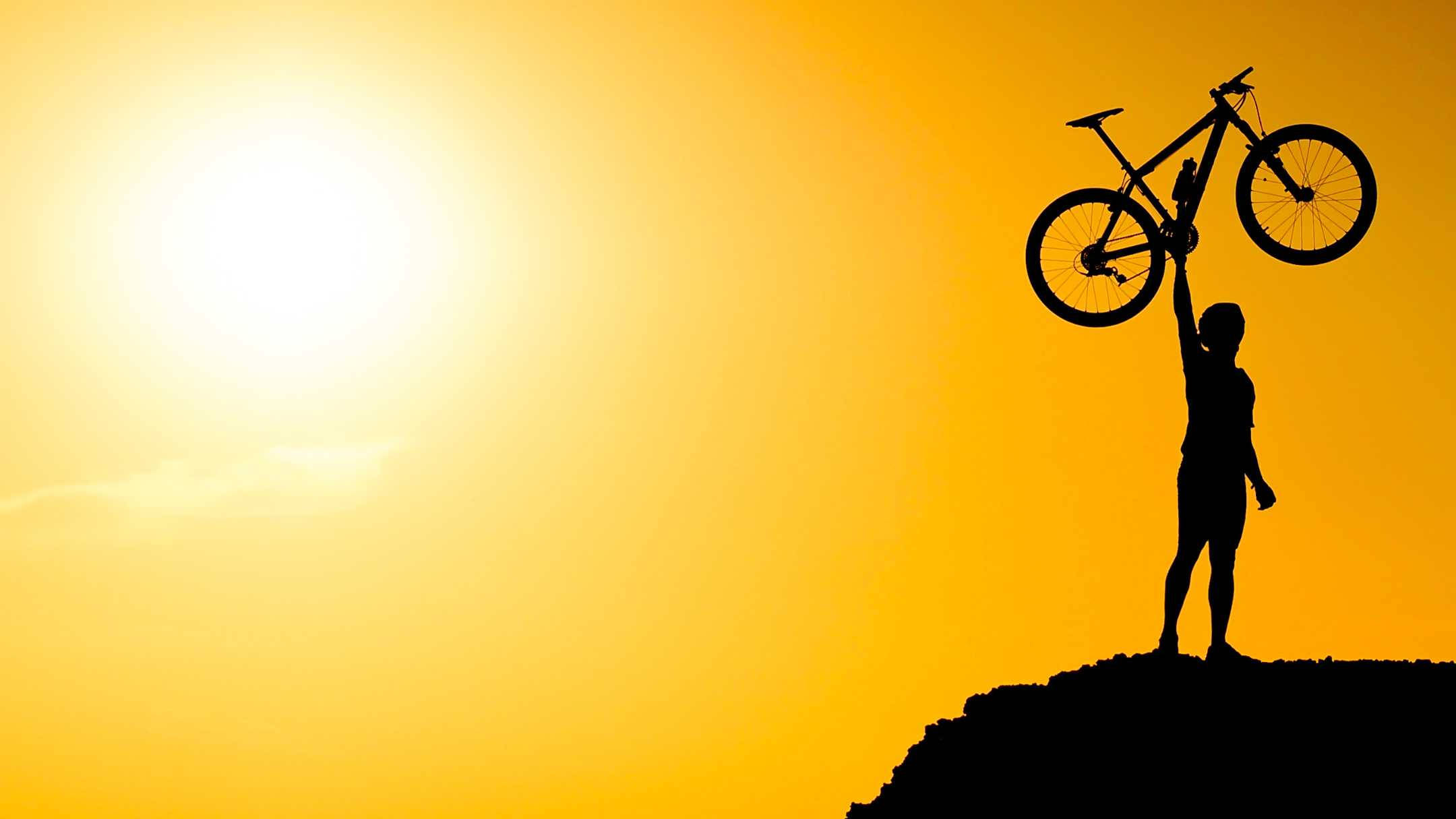 Mountain Bike 桌布 壁紙 — 金色夕陽下，一名自行車手在懸崖頂上勝利地高舉登山車的剪影。