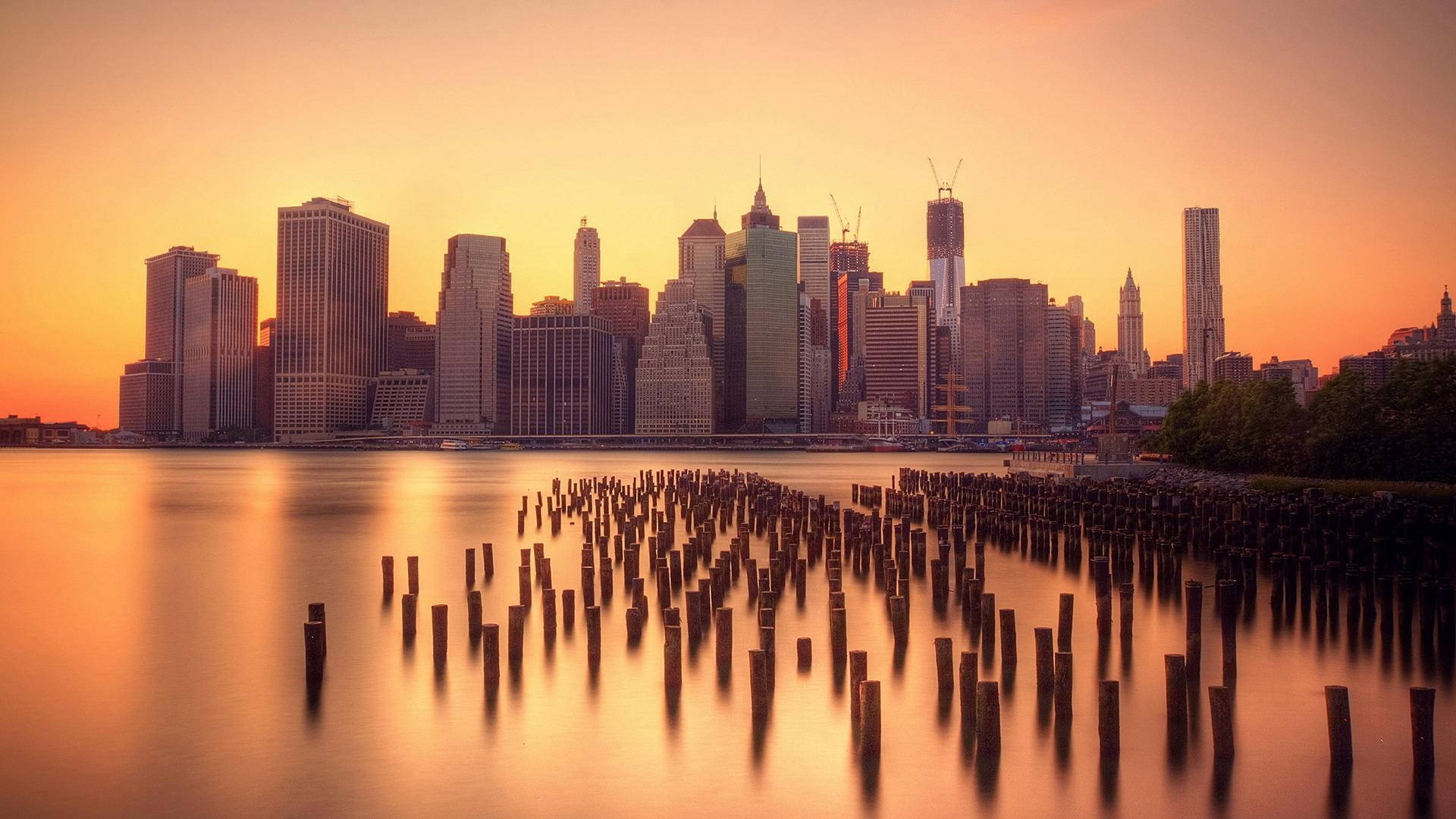 manhattan 桌布 壁紙 — 金色夕陽以溫暖光線沐浴紐約天際線，平靜水面上倒映著光芒，前景有風化的木樁。