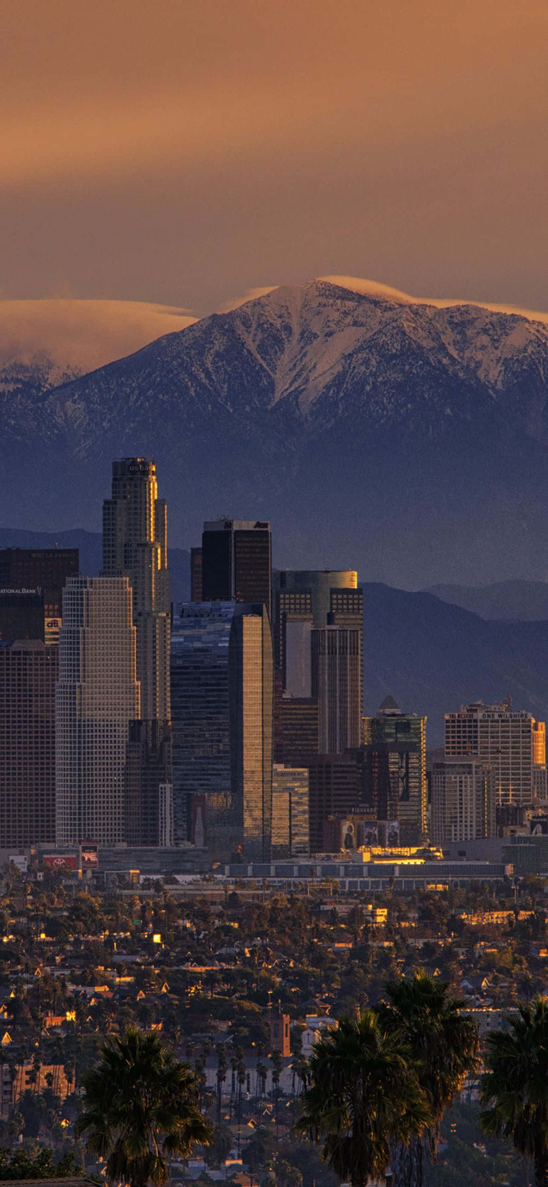 iPhone Xs California 桌布 壁紙 — 金色夕陽以溫暖光線沐浴著市中心的摩天大樓和棕櫚樹，背後是白雪覆蓋的山脈。