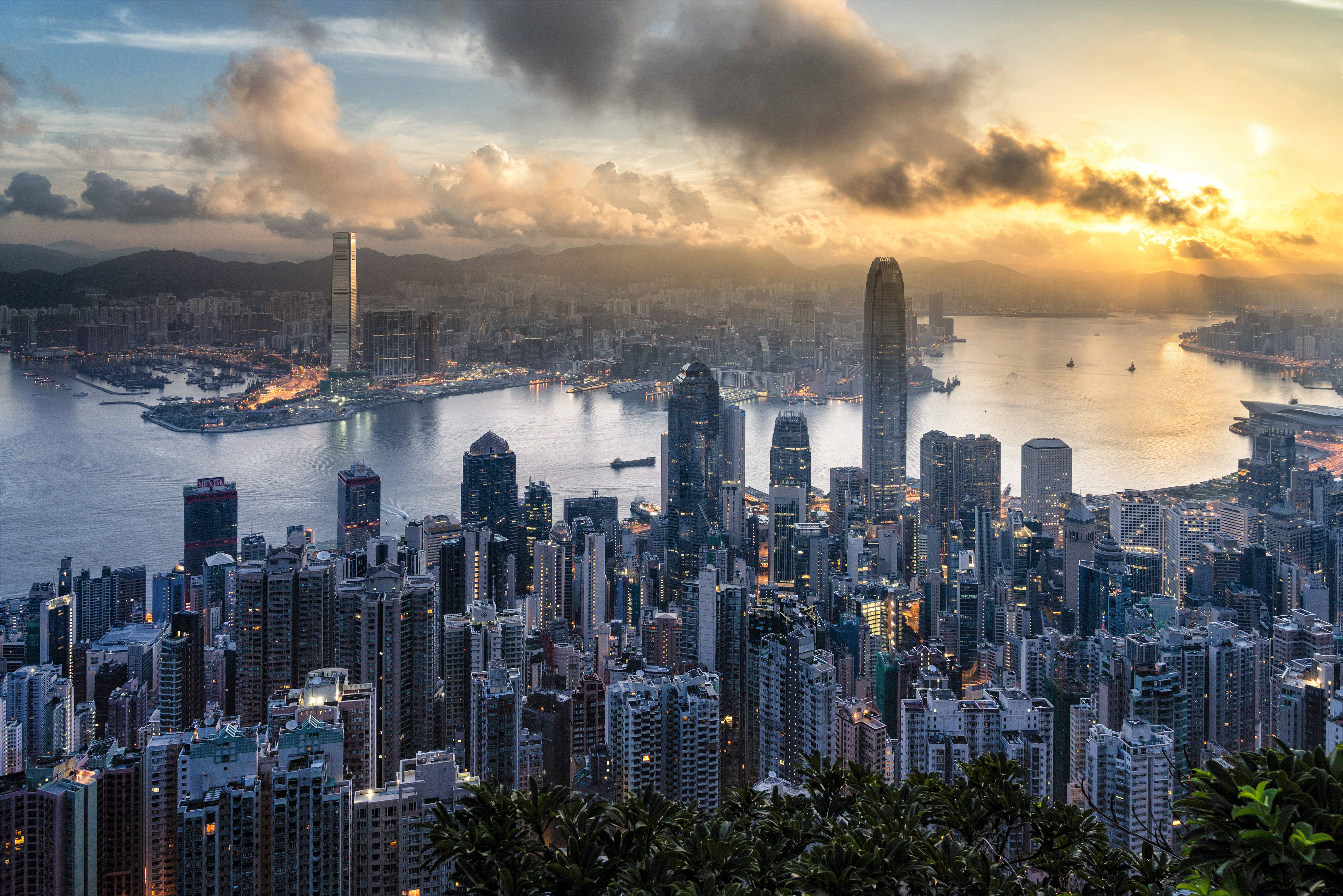 香港 桌布 壁紙 — 金色夕陽以溫暖光線沐浴香港天際線，雲朵飄過港口，高聳摩天大樓反射逐漸消退的光芒。