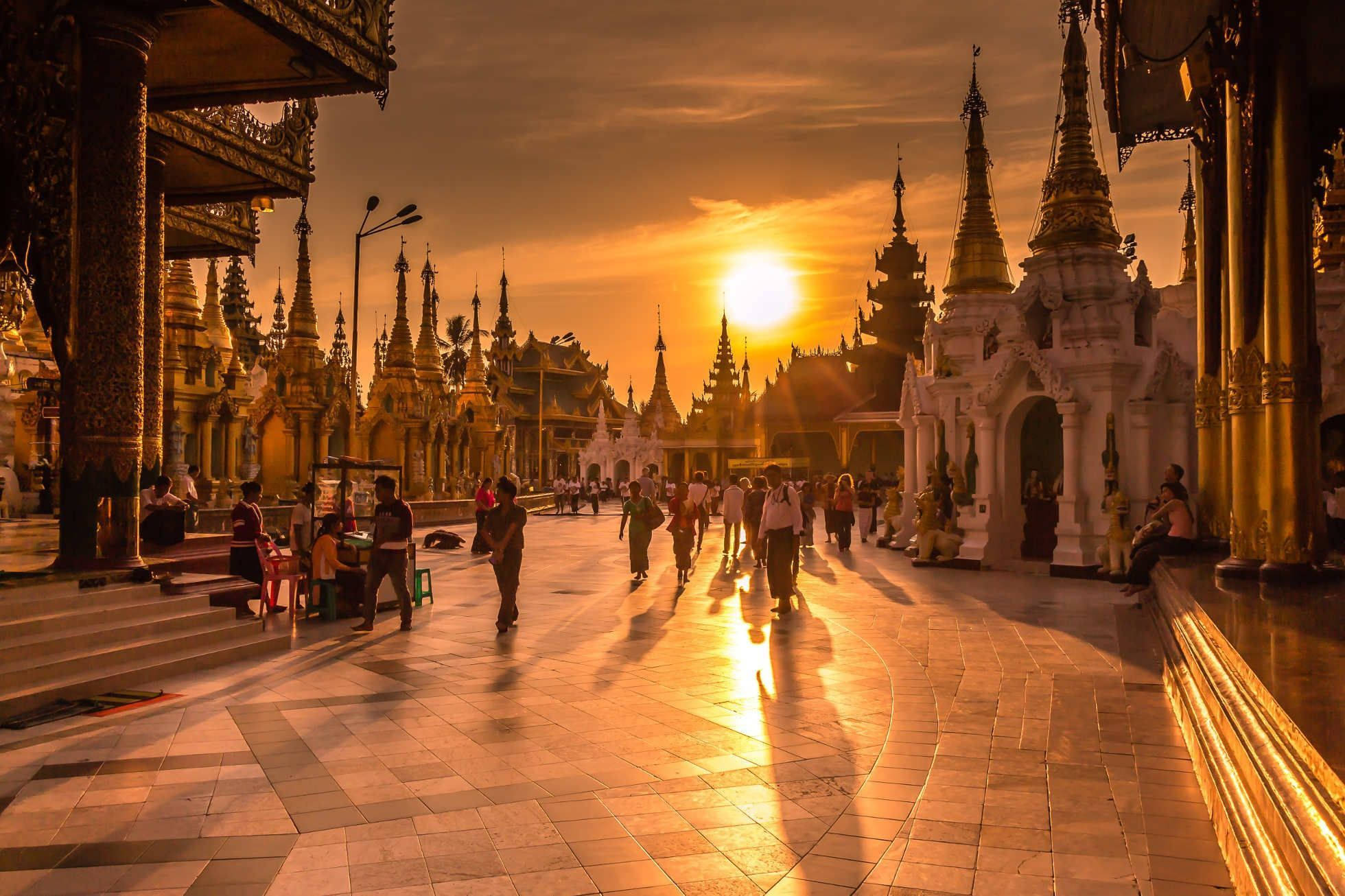 Yangon 桌布 壁紙 — 金色夕陽以溫暖光芒沐浴著熙攘的寺廟庭院，遊客漫步經過華麗的尖塔和鍍金柱子。