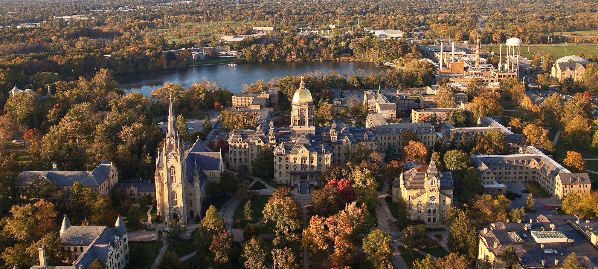 Notre Dame 桌布 壁紙 — 金色夕陽光下，被秋葉和反射湖面環繞的宏偉大學校園鳥瞰圖。