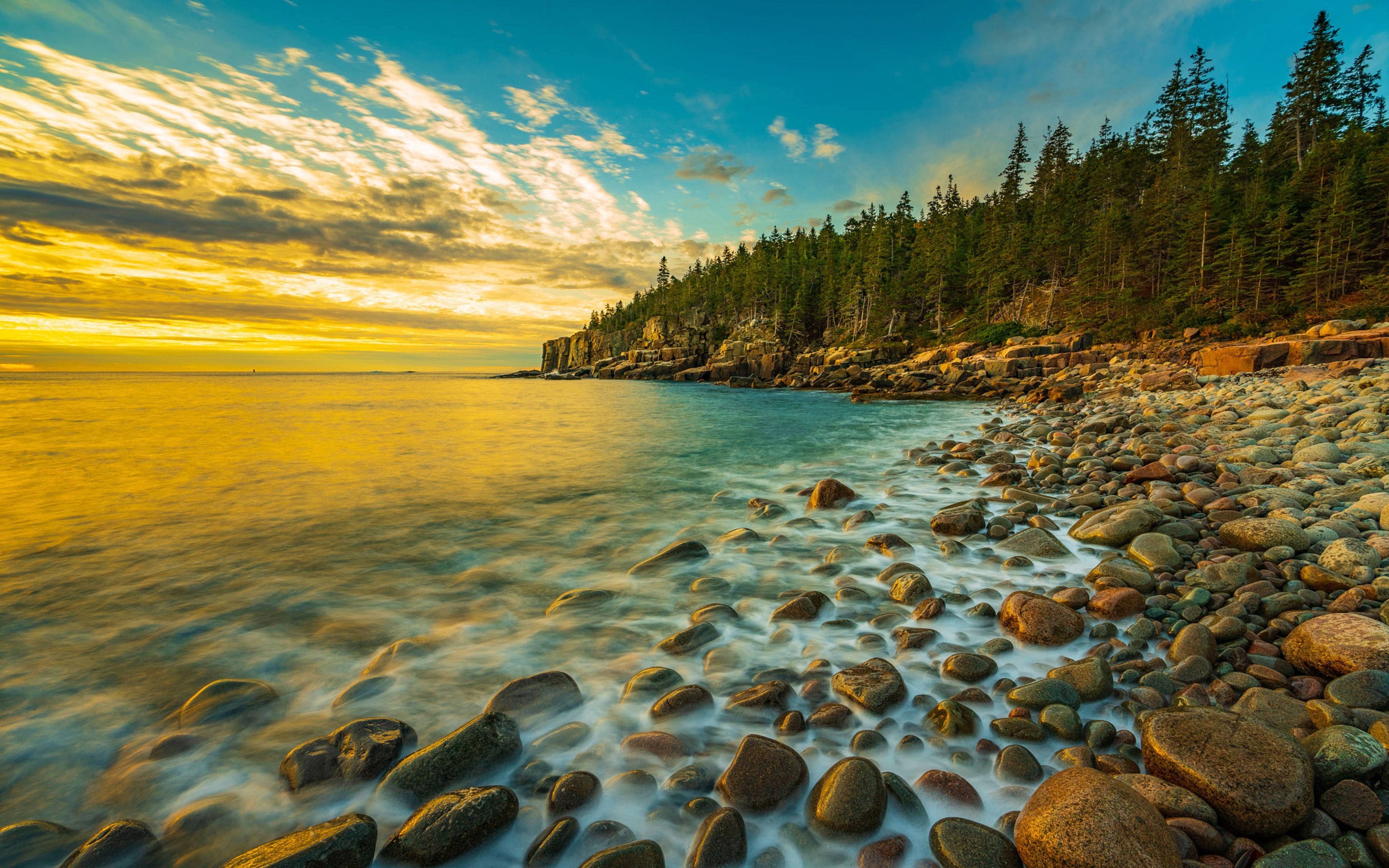 Stone 桌布 壁紙 — 金色夕陽在平靜的海浪上閃耀，海浪沖刷著長滿常綠樹的岩石海岸。