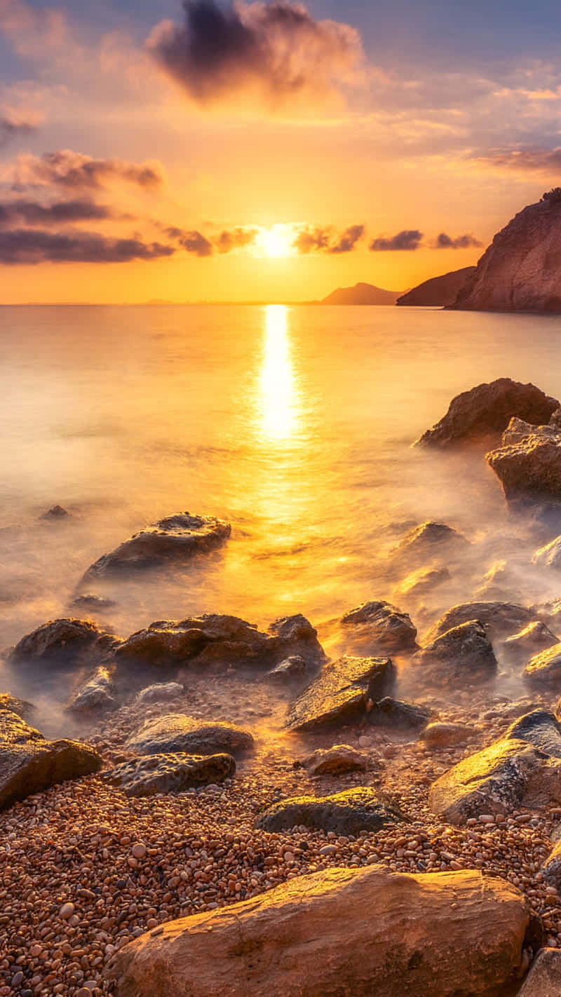 Sunrise iPhone 桌布 壁紙 — 金色夕陽在平靜的海面上閃耀，光線映照在霧氣籠罩的岩石和卵石灘上。