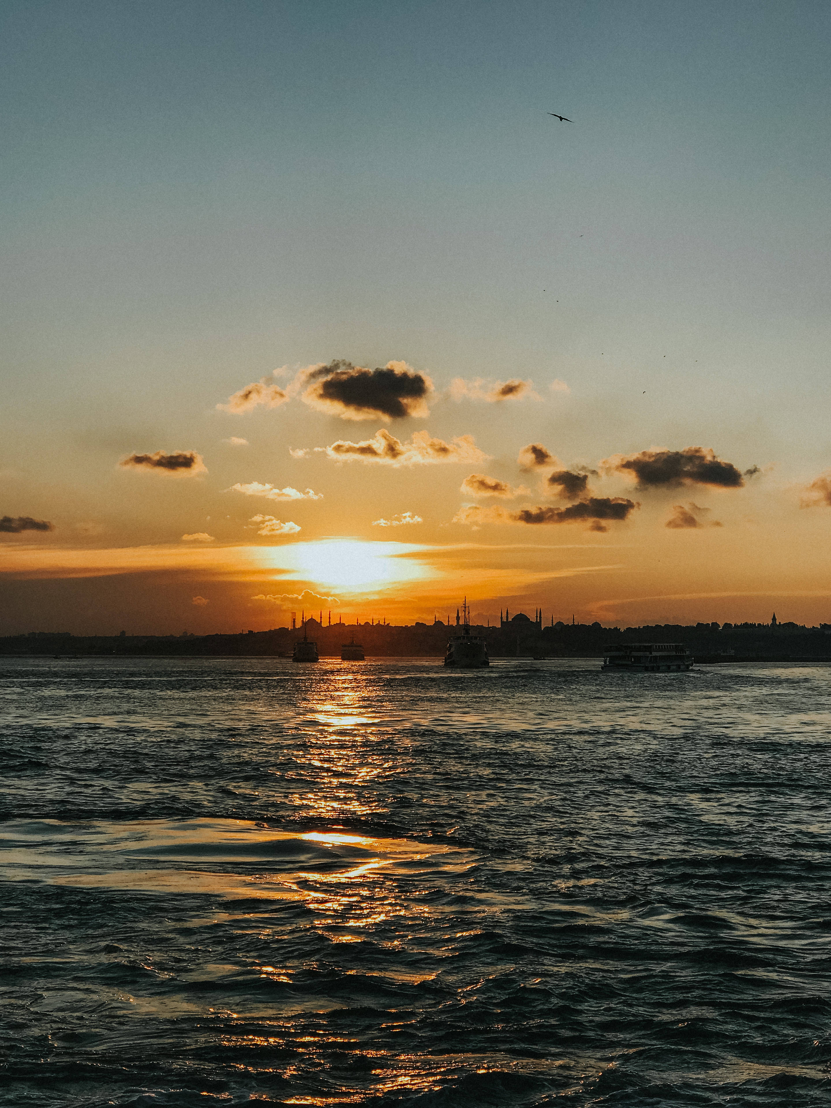 日落 桌布 壁紙 — 金色夕陽在水面上閃耀，船隻剪影和遠處城市天際線在散落的雲層下。