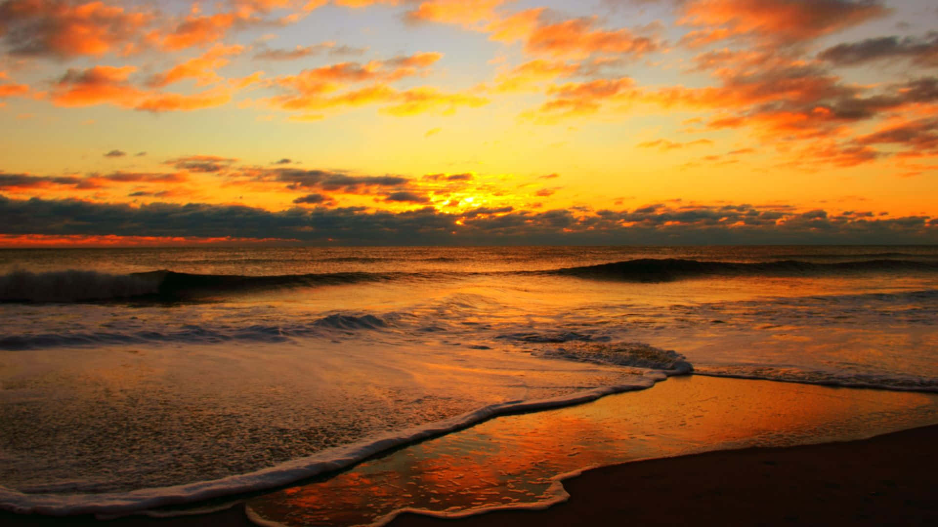 Beach 壁紙 — 金色夕陽將海洋和天空染成火熱的橙色和琥珀色，海浪拍打著深色沙灘。