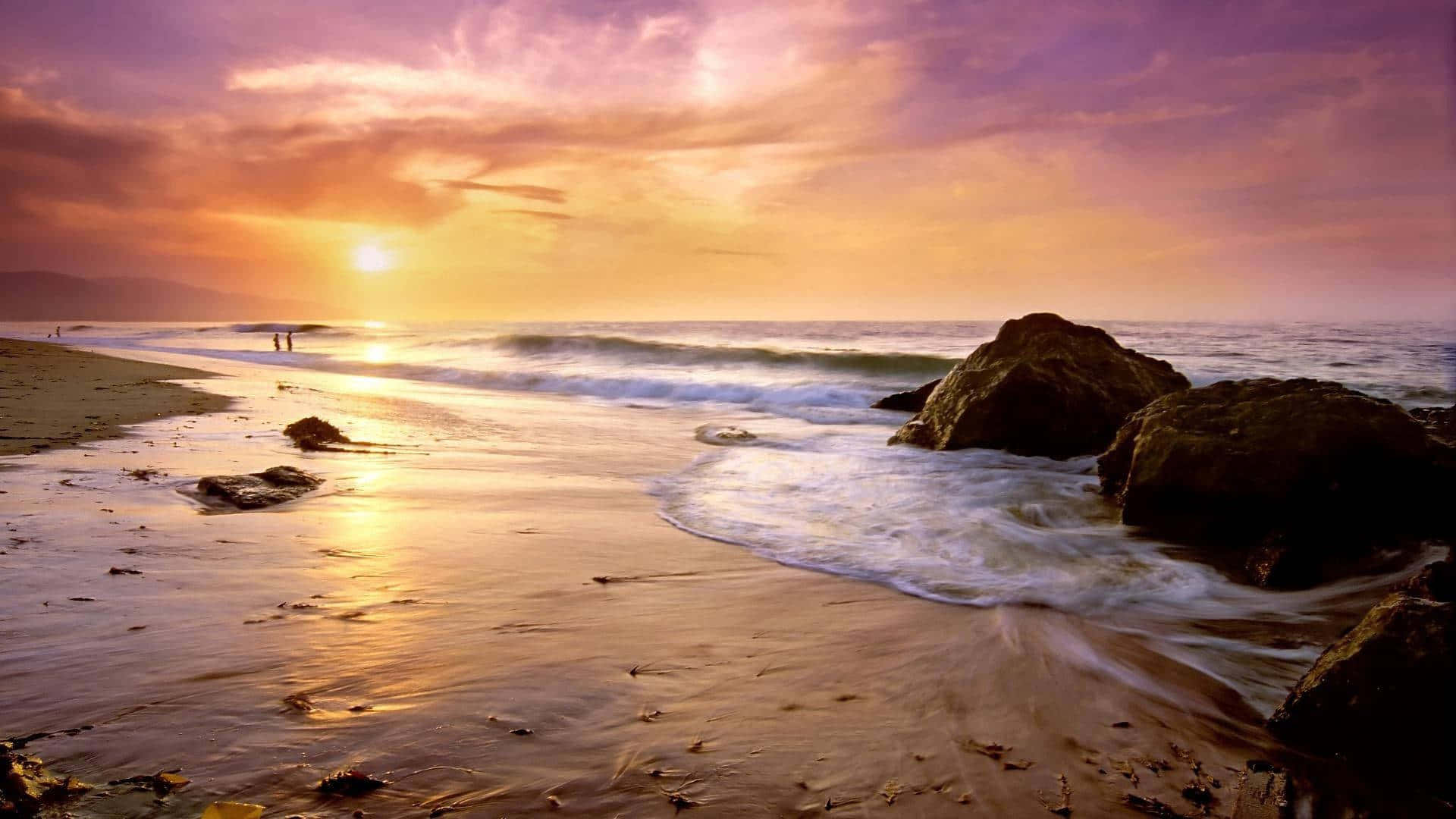 1920x1080 Malibu 桌布 壁紙 — 金色夕陽將海洋和沙灘染上溫暖色調，海浪拍打著暗色岩石，兩個人影沿著海岸漫步。