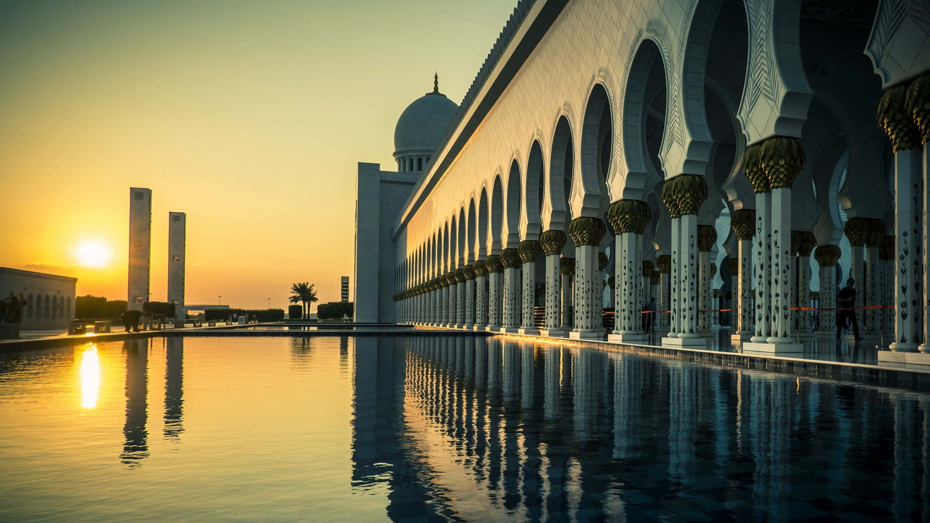 Uae 桌布 壁紙 — 金色夕陽將溫暖光線投射在宏偉清真寺拱門上，倒映在平靜的水面。