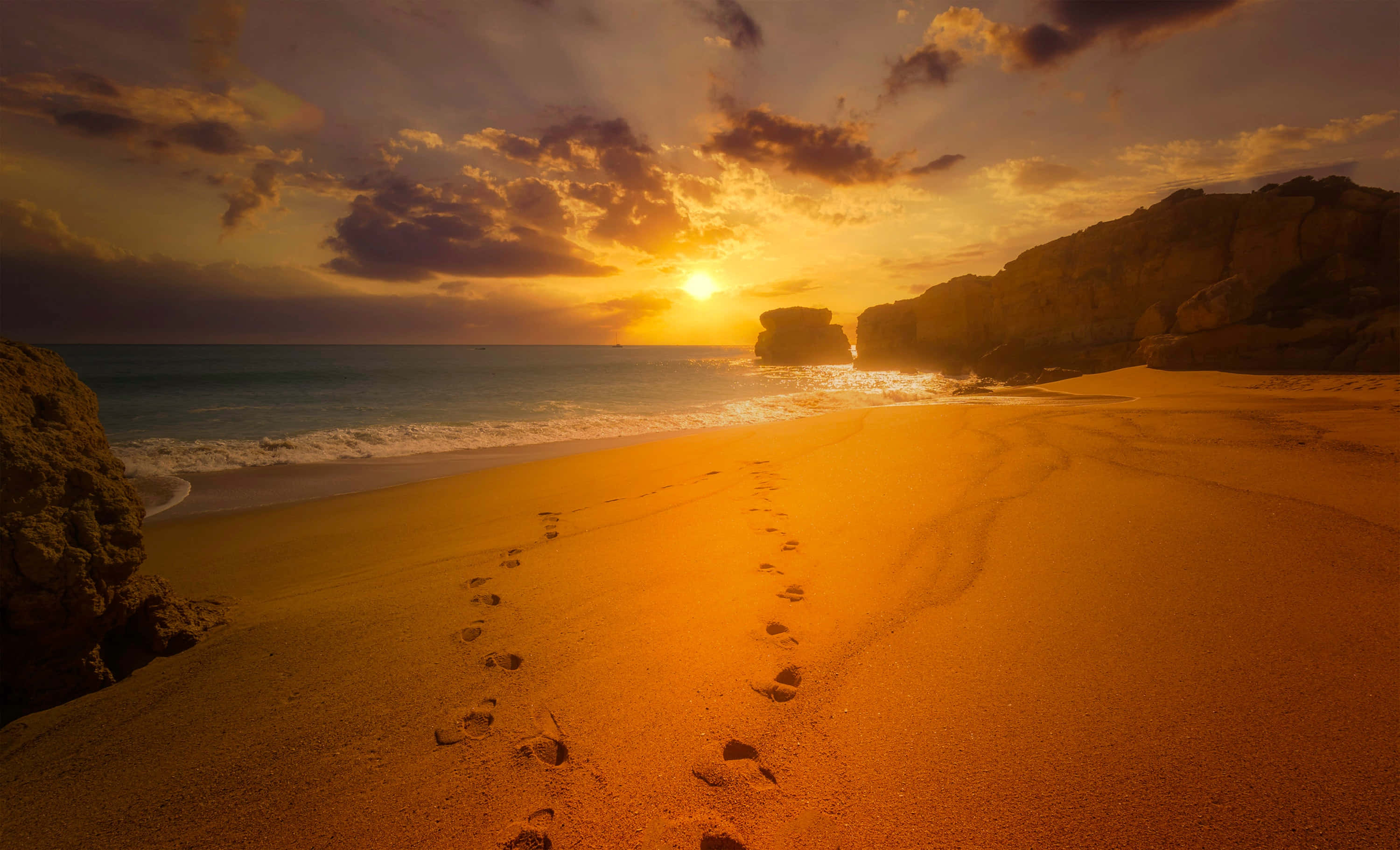 Seashore 桌布 壁紙 — 金色夕陽將溫暖的光線投射在沙灘上，腳印通向海洋和岩石峭壁。