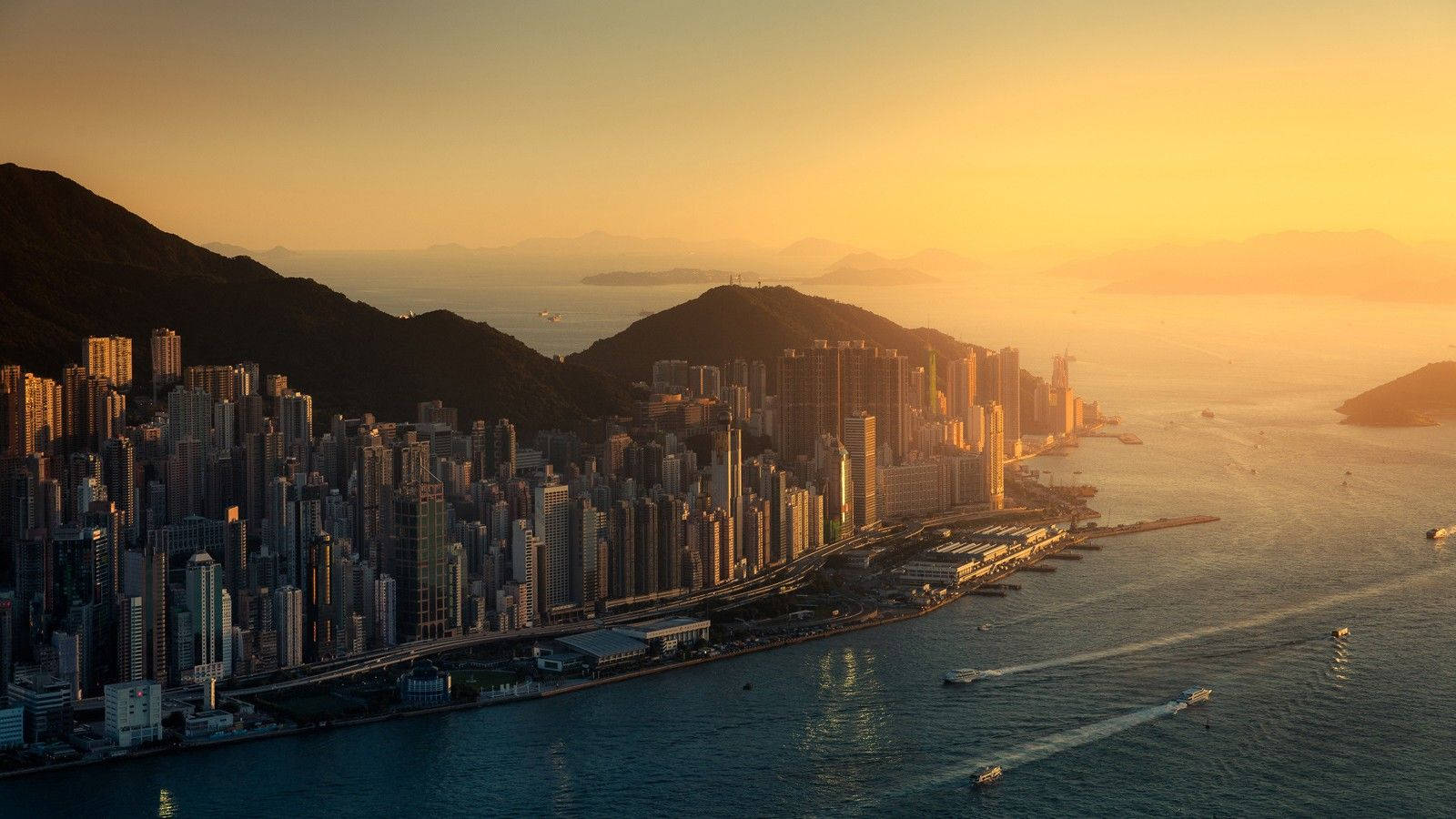城市風景 桌布 壁紙 — 金色夕陽將香港天際線沐浴在溫暖的光芒中，船隻在港口水面上滑行。