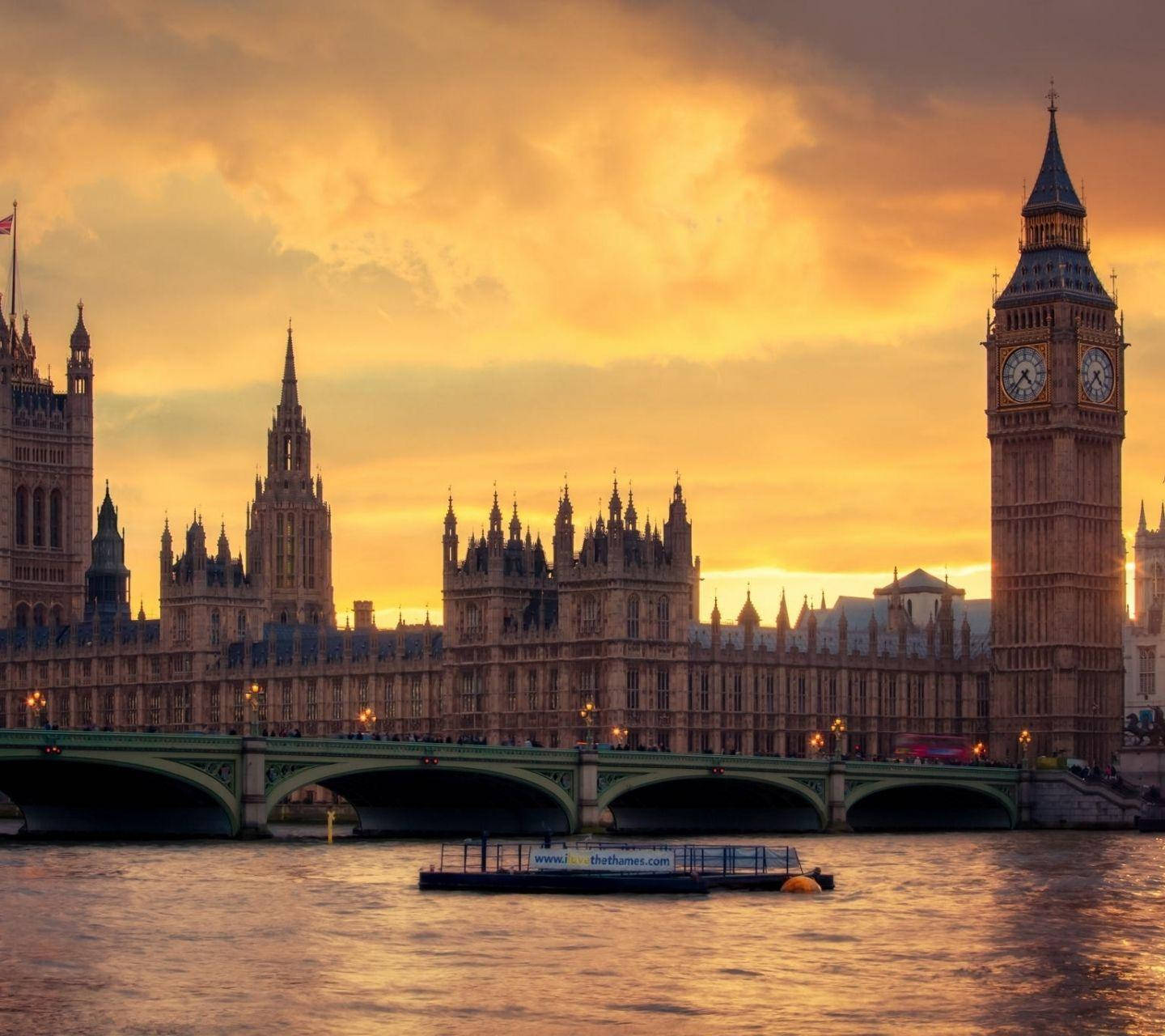 England 桌布 壁紙 — 金色夕陽沐浴倫敦標誌性大笨鐘和國會大廈，溫暖光線中，一艘河船在威斯敏斯特橋下滑行。