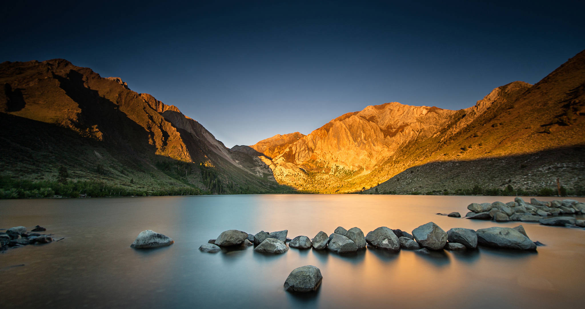 加州 桌布 壁紙 — 金色夕陽沐浴崎嶇山頂，溫暖光線反射在平靜湖面，前景散落岩石。