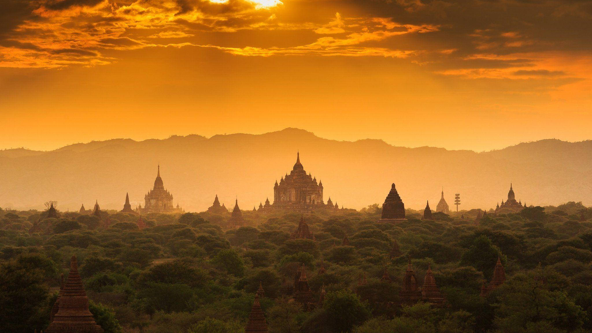 Burma 桌布 壁紙 — 金色夕陽沐浴著古老的寶塔，溫暖的光芒中，它們從綠樹成蔭的海洋中升起，映襯著遠山。