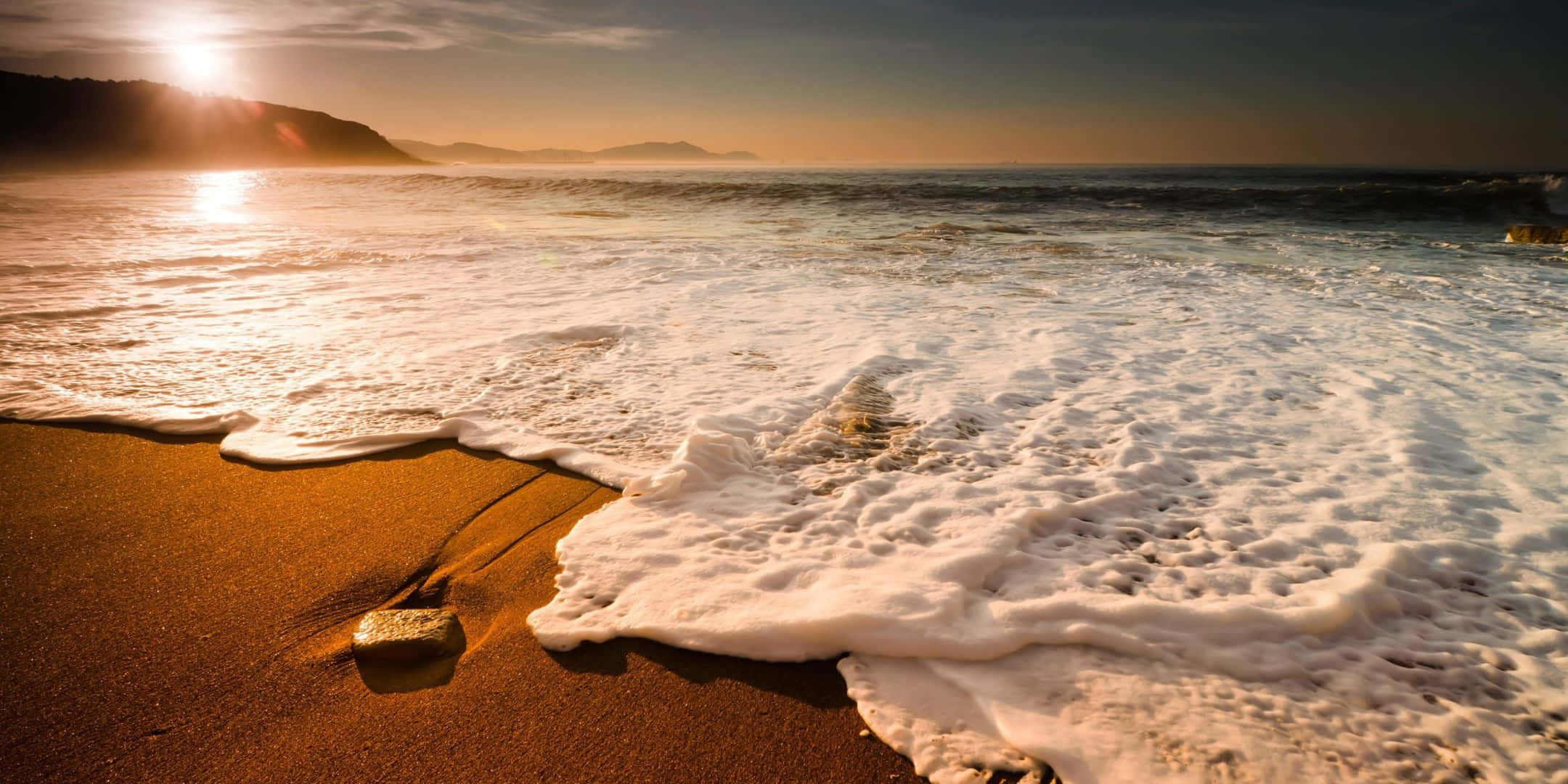 2160x1080 桌布 壁紙 — 金色夕陽海浪拍打溫暖沙灘，帶有泡沫白色浪尖，發光的太陽倒映在水面上。