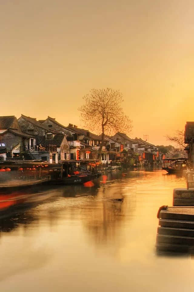 日落 桌布 壁紙 — 金色夕陽灑落在寧靜的運河小鎮，傳統房屋和停泊的船隻在平靜的水面上溫暖地倒映。