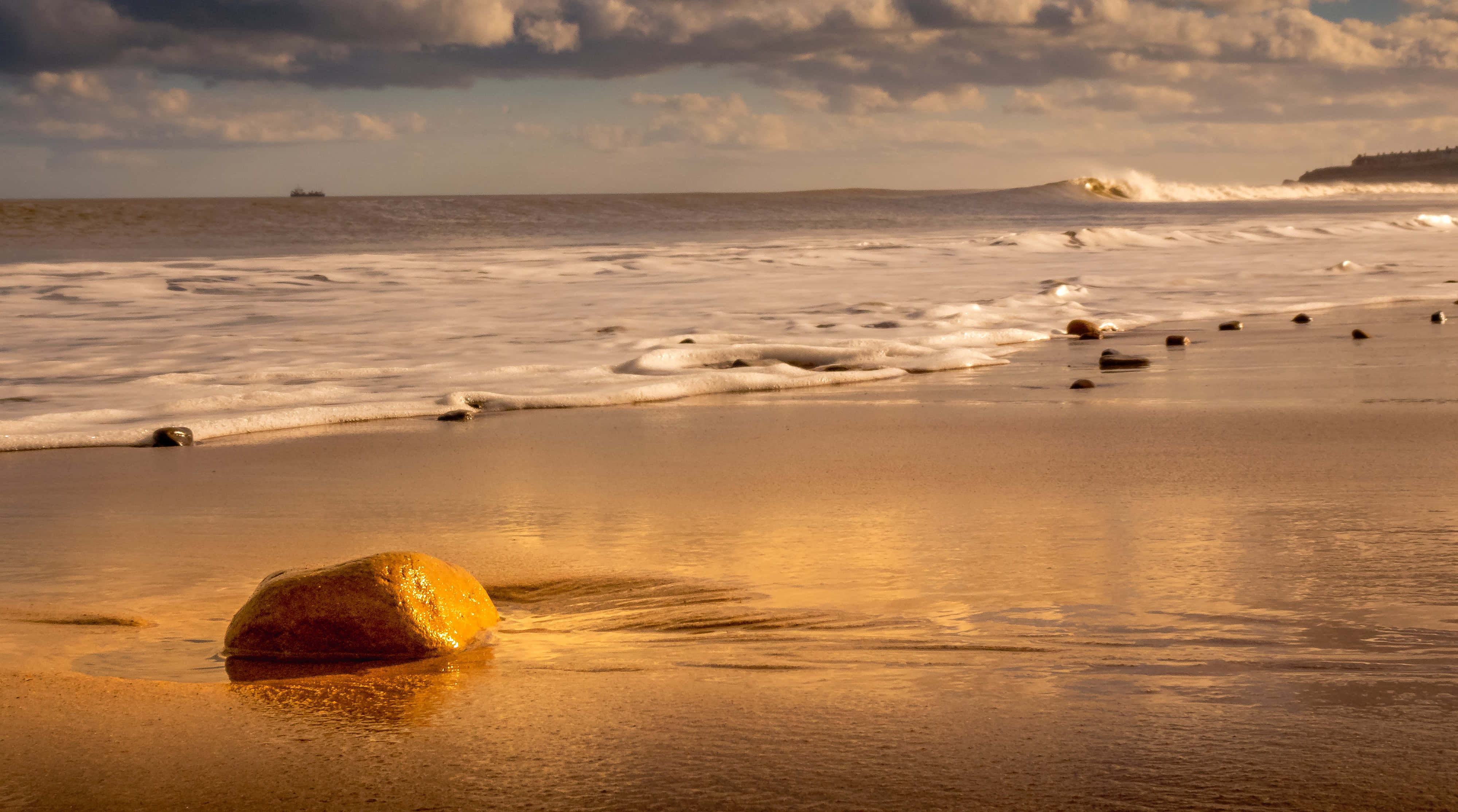 Aesthetic Beachy 桌布 壁紙 — 金色夕陽灑落在濕潤的沙灘上，海浪拍打，一塊光滑的大石在前景閃爍。