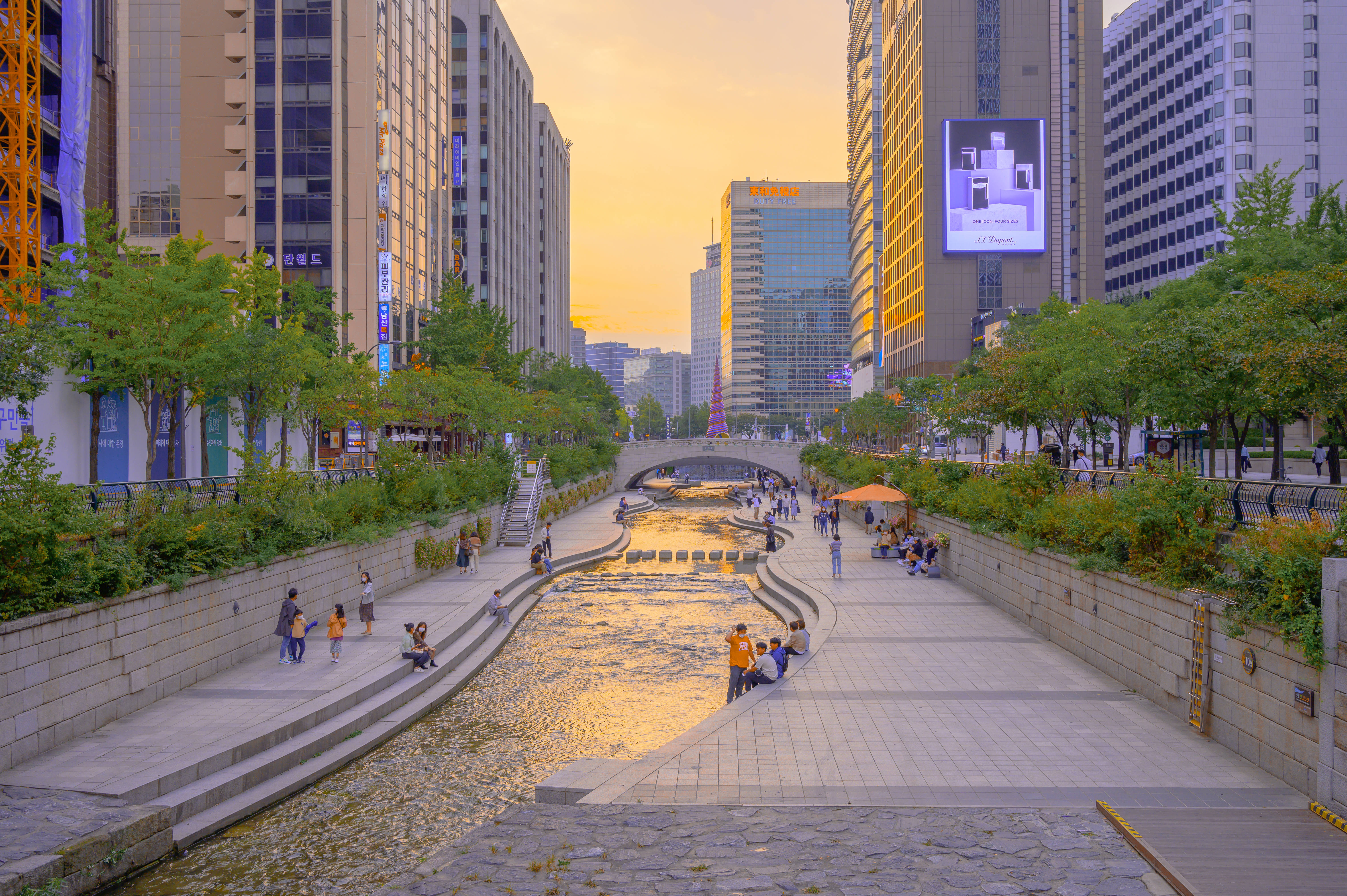 首爾 桌布 壁紙 — 金色夕陽灑落在繁忙的城市運河，兩旁樹木和現代摩天大樓林立，人們在石徑上放鬆。