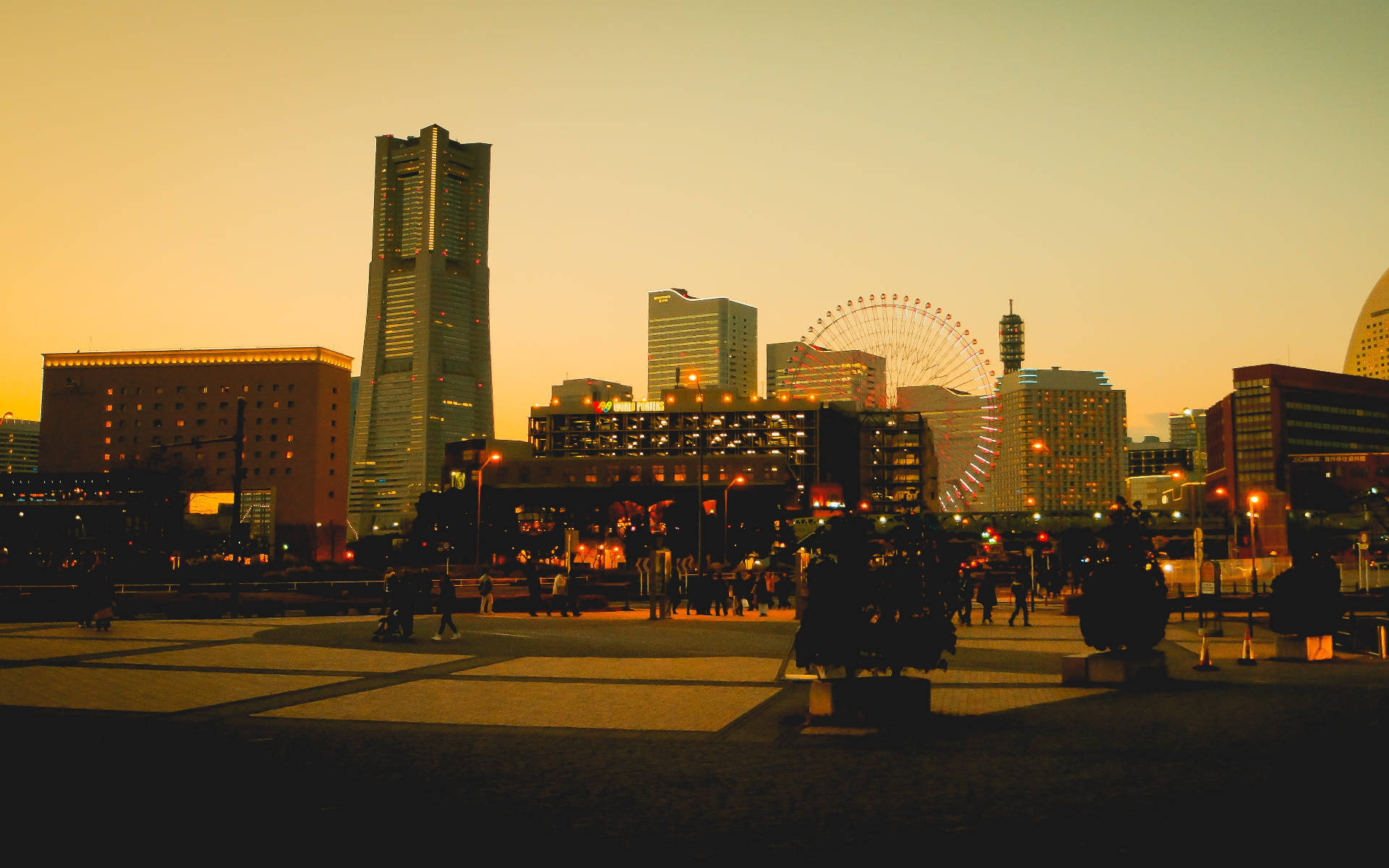 橫濱 桌布 壁紙 — 金色夕陽灑落在繁華的城市廣場上，映襯著人們的剪影和包括摩天輪在內的宏偉建築。