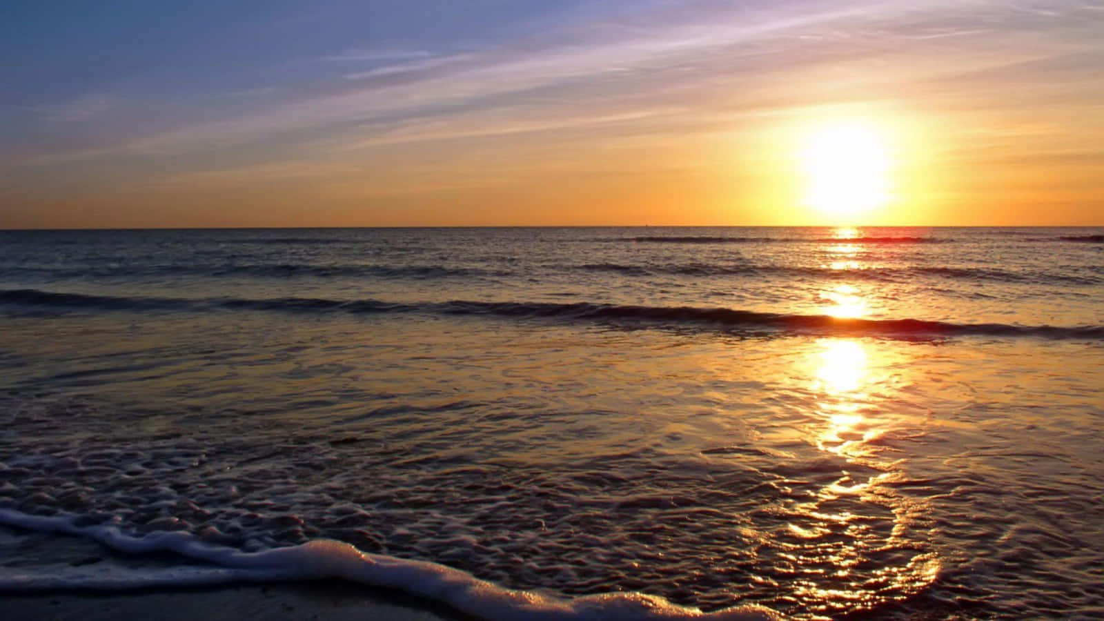 日落 桌布 壁紙 — 金色夕陽照耀在平靜的海浪上，沖刷著沙灘，泛起柔軟的泡沫。