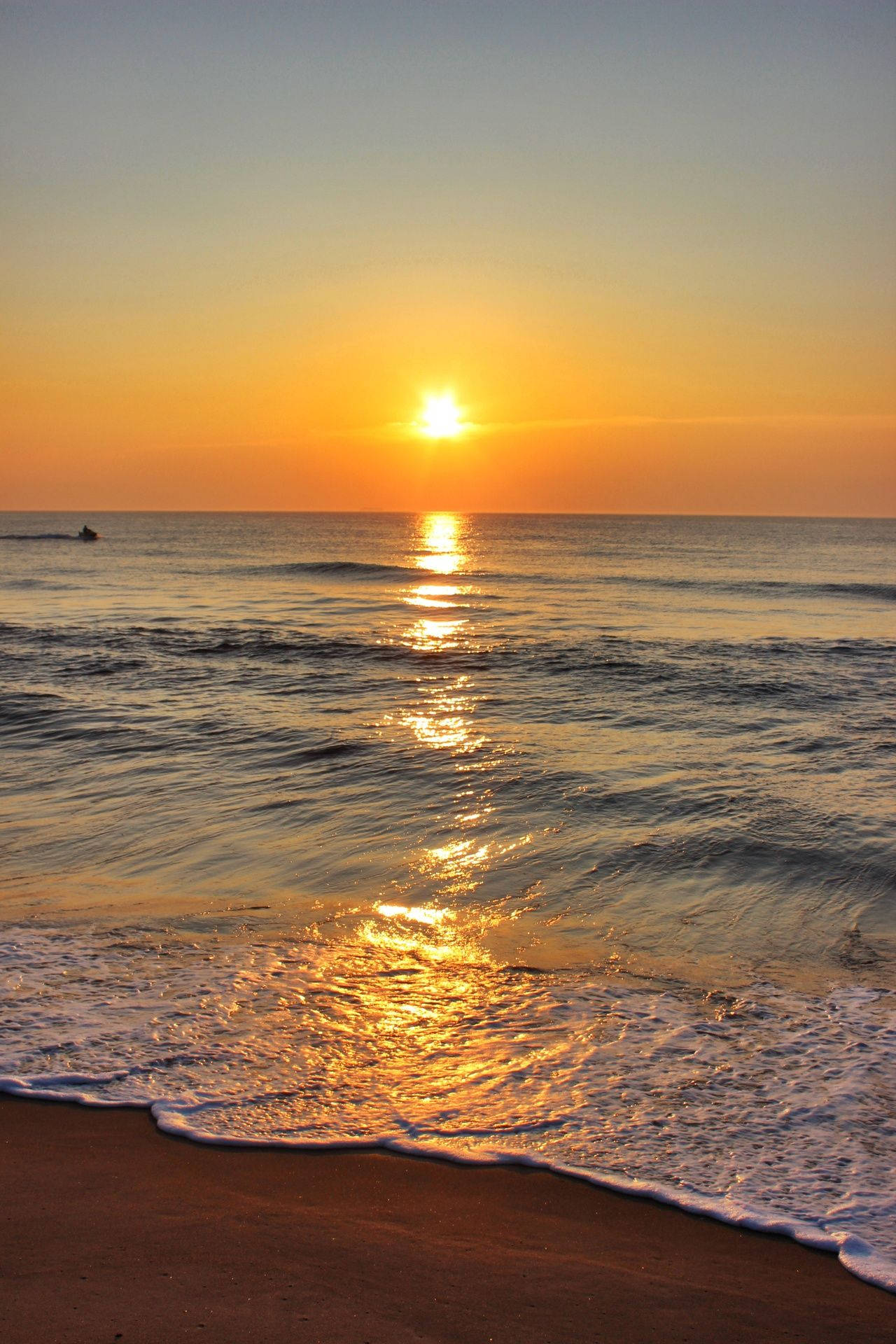 海洋 桌布 壁紙 — 金色夕陽照耀在平靜的海浪上，沖刷著沙灘，遠處有游泳者。
