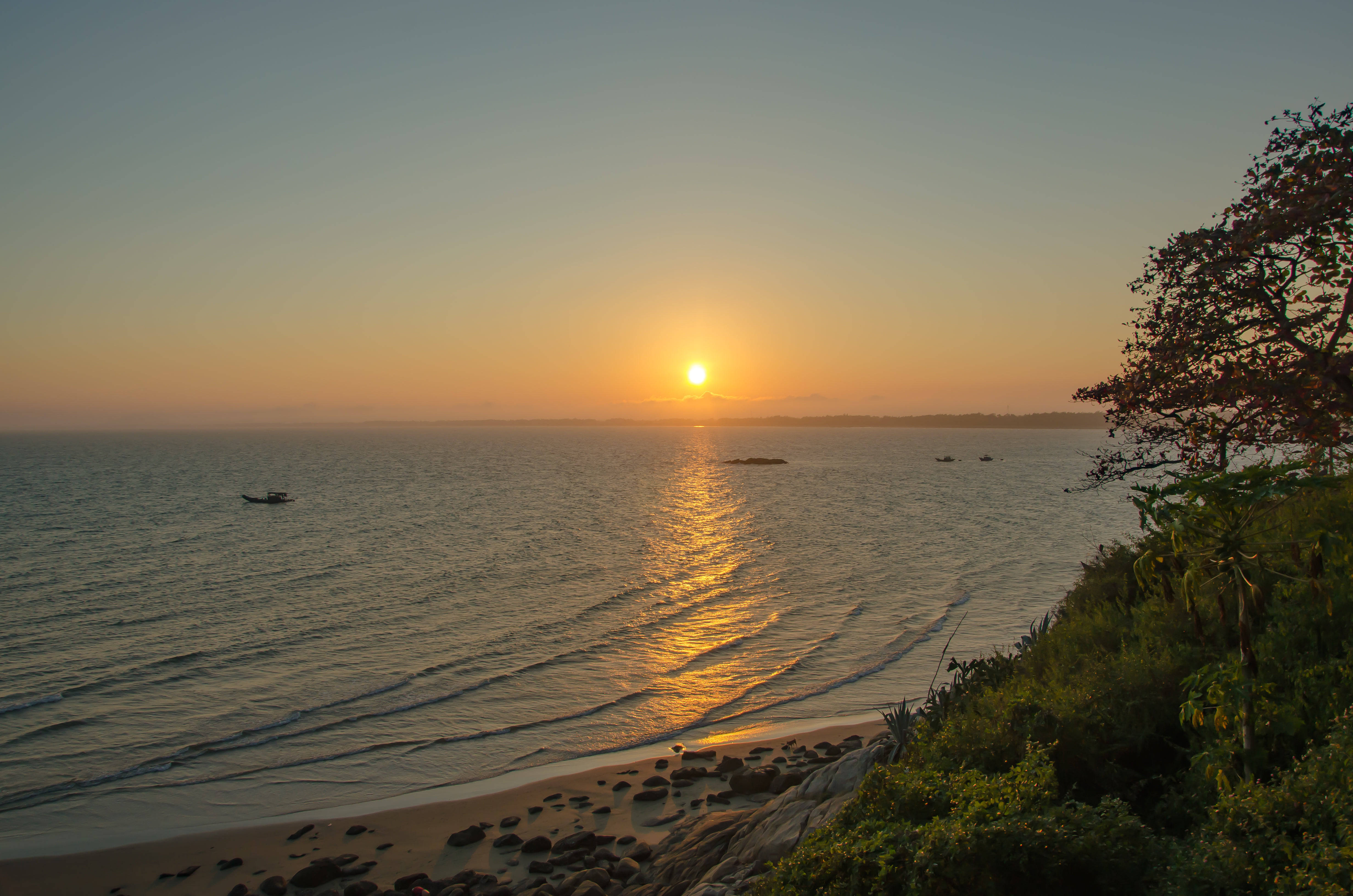 Calming 桌布 壁紙 — 金色夕陽照耀在平靜的海面上，小船在綠意盎然的岩石海岸附近漂流。