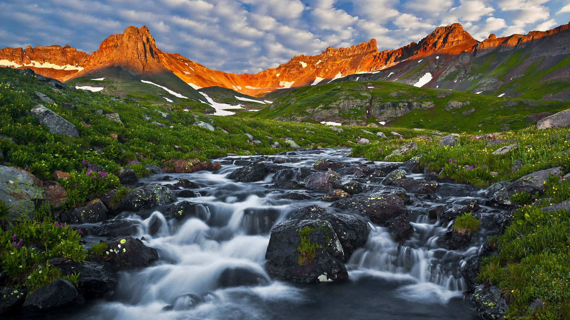 Colorado Mountains 桌布 壁紙 — 金色夕陽照耀著崎嶇的山峰，一條奔流的小溪穿過鮮綠的草地，衝過黑色的岩石。