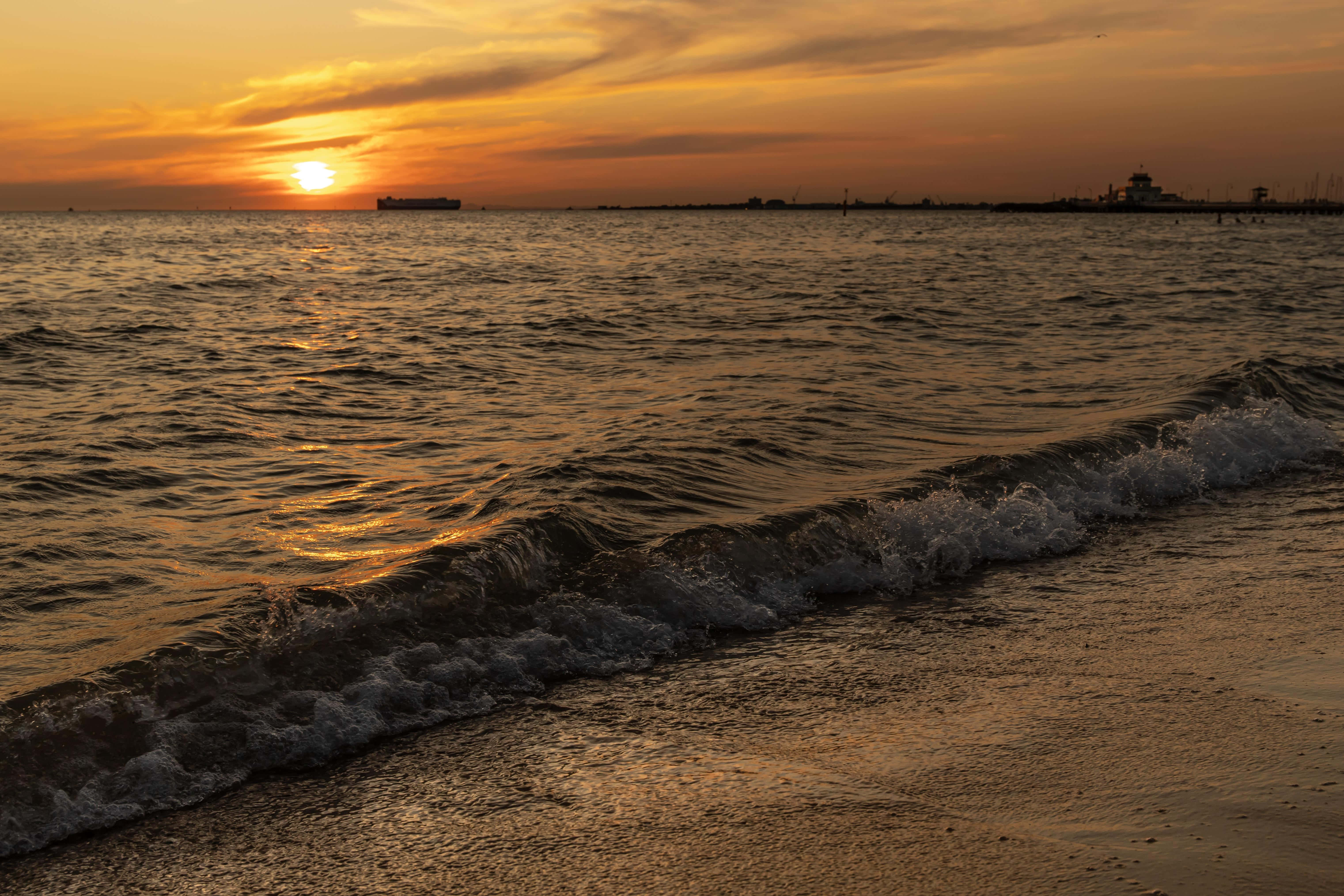 海灘 桌布 壁紙 — 金色夕陽照耀著海洋波浪拍打沙灘，遠處有船和燈塔的剪影。