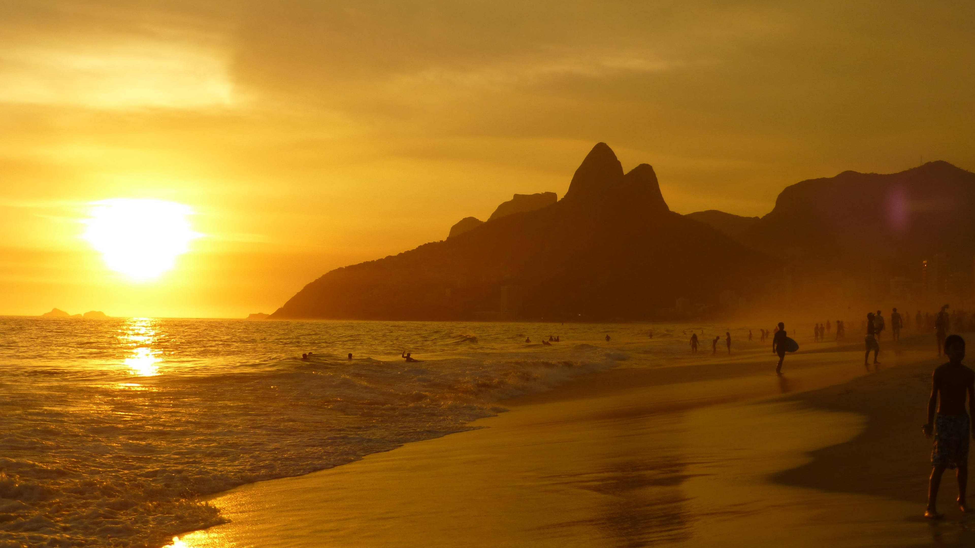 rio 桌布 壁紙 — 金色夕陽照耀著熱鬧的海灘，遠處有剪影人物和標誌性的山峰。