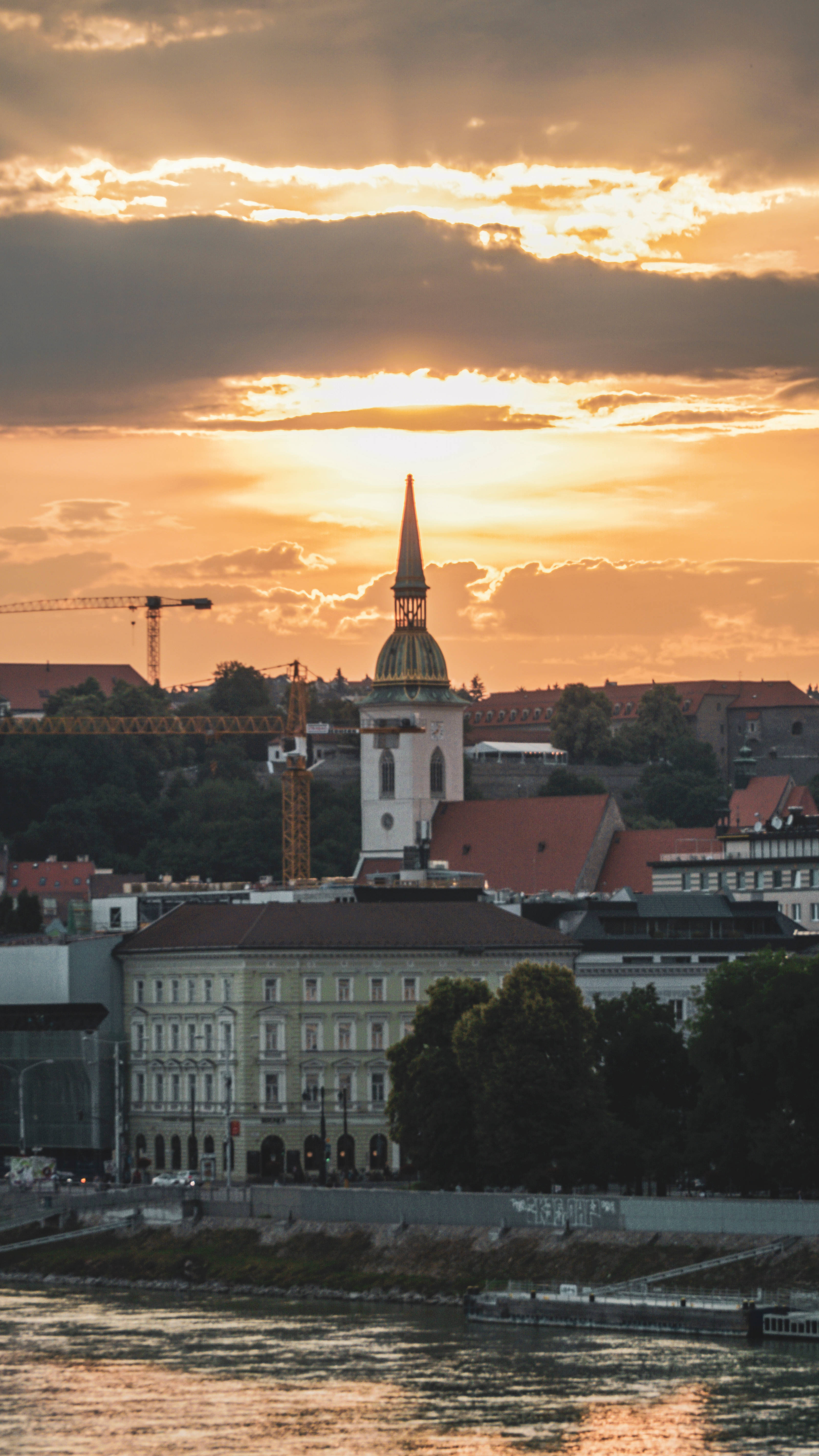 Slovakia 桌布 壁紙 — 金色夕陽穿過雲層，照耀在歷史悠久的教堂尖頂和河畔建築上。