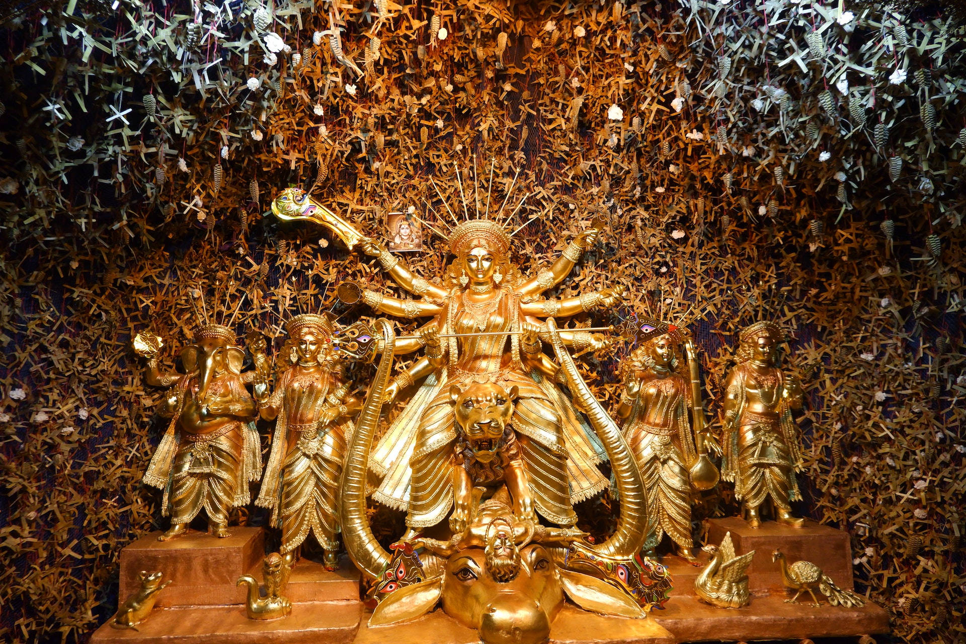 Hindu God 桌布 壁紙 — 金色多臂多武器的杜爾迦雕像，周圍環繞著較小的金色神祇，背景是懸掛的紙鶴。