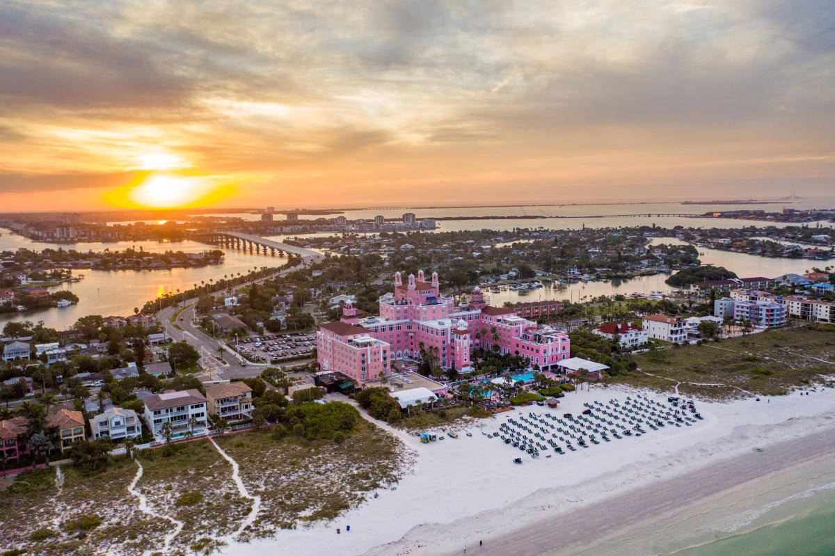 Florida Beach 桌布 壁紙 — 金色天空下，白色沙灘和寧靜水域旁的鮮豔粉紅色酒店的鳥瞰圖。