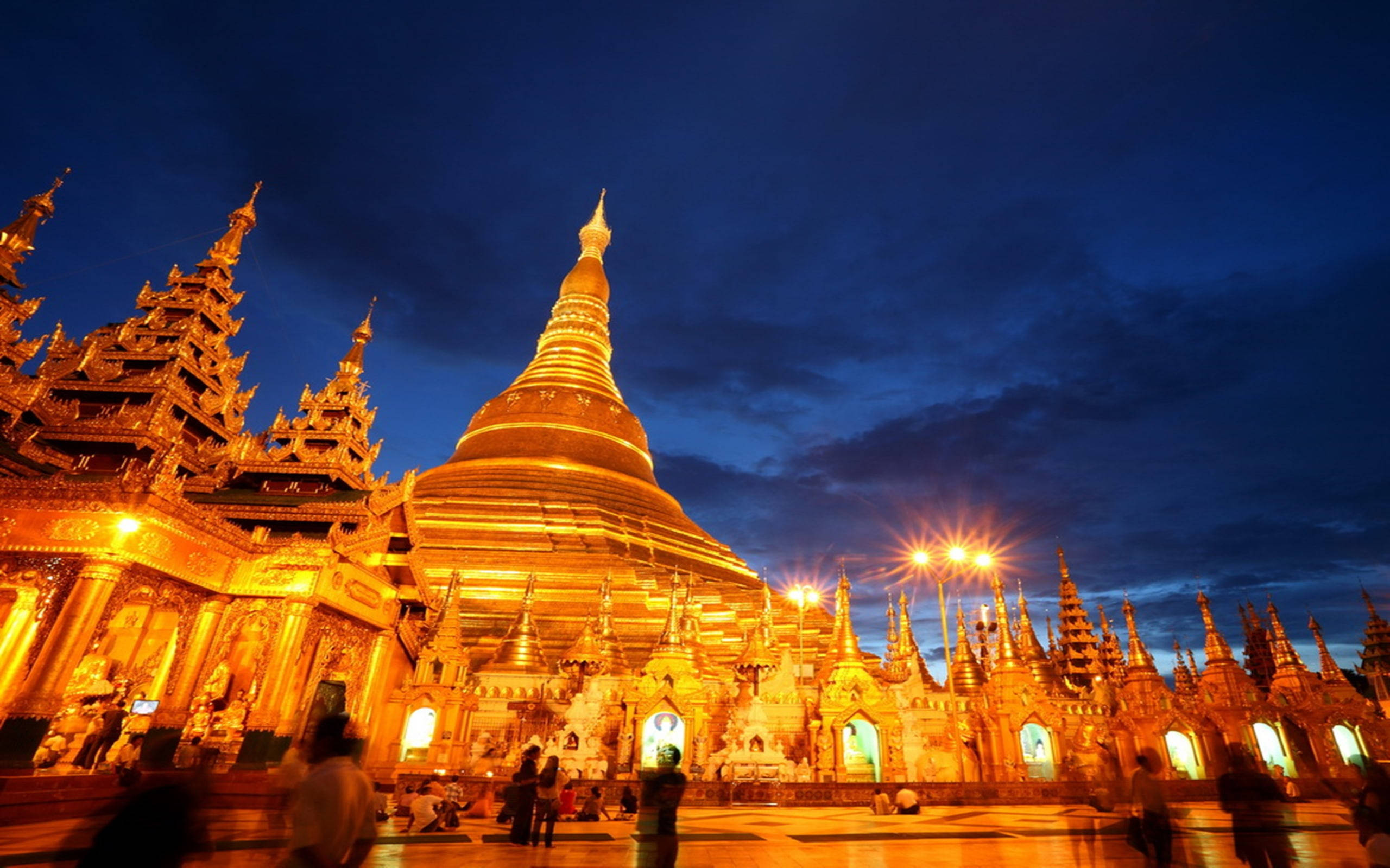 Yangon 桌布 壁紙 — 金色寶塔在黃昏時亮起，發光的尖頂映襯著深藍色的天空，下方有模糊的遊客。