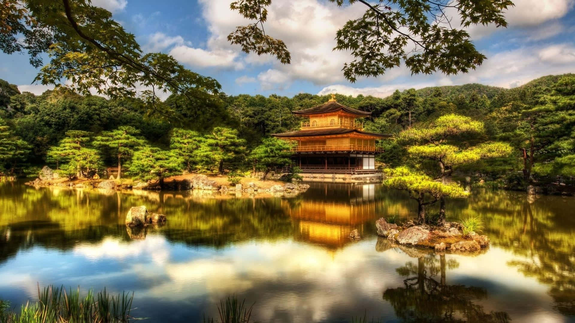 禪 桌布 壁紙 — 金色寺廟亭閣倒映在平靜的水面上，周圍是茂密的綠樹，頭頂是明亮的藍天。