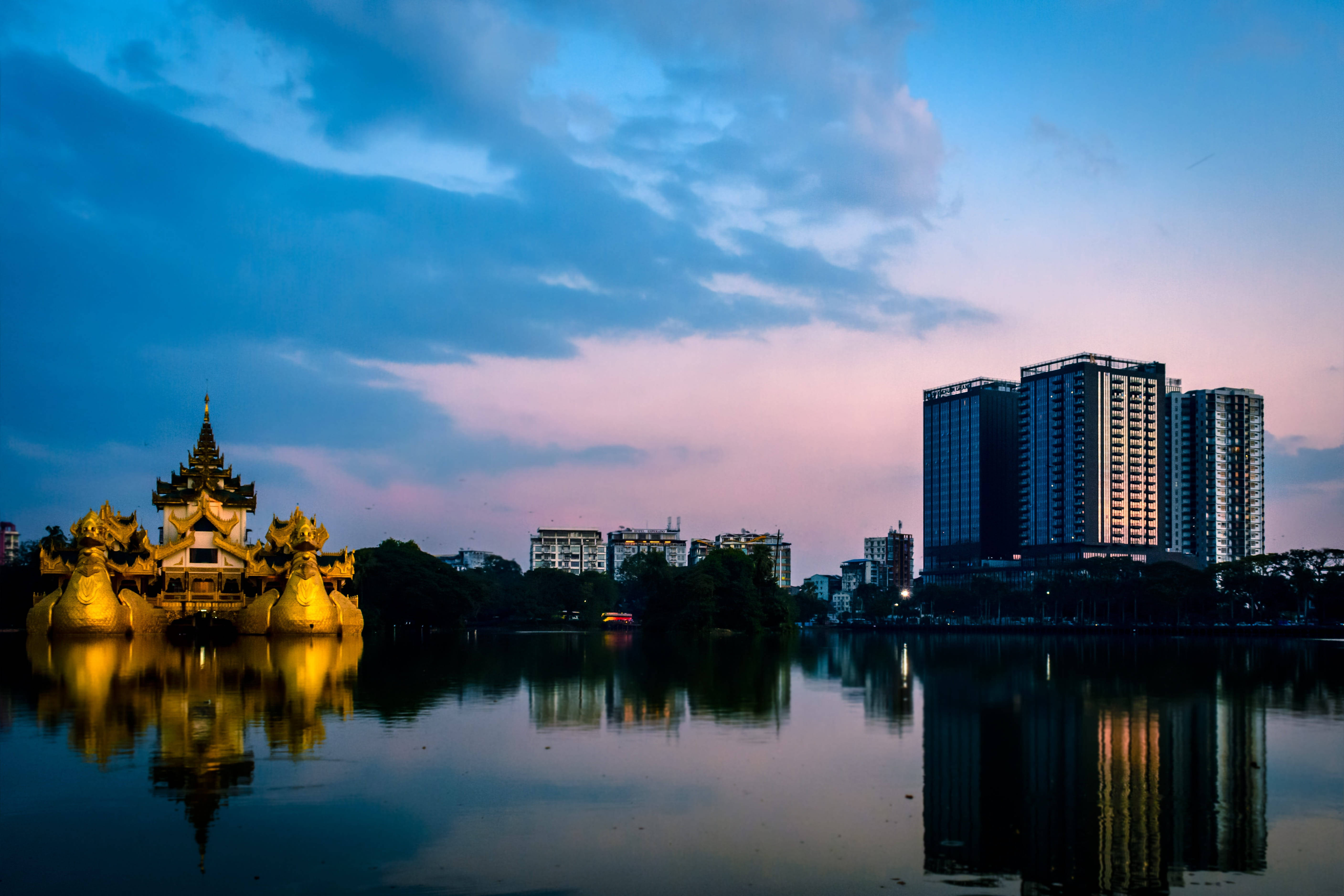 Yangon 桌布 壁紙 — 金色寺廟倒映在平靜的水面上，現代摩天大樓在染上粉紅和藍色調的暮色天空中聳立。