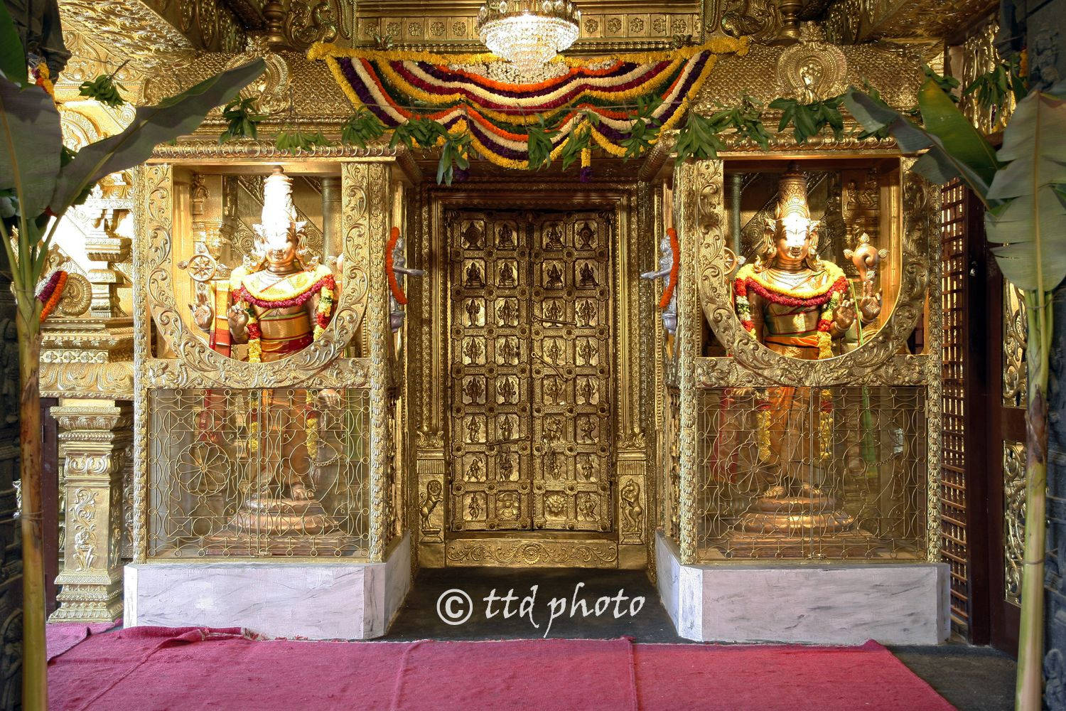 Hindi 桌布 壁紙 — 金色寺廟入口，兩側有兩尊華麗的神像，上方掛著彩色花環和吊燈。