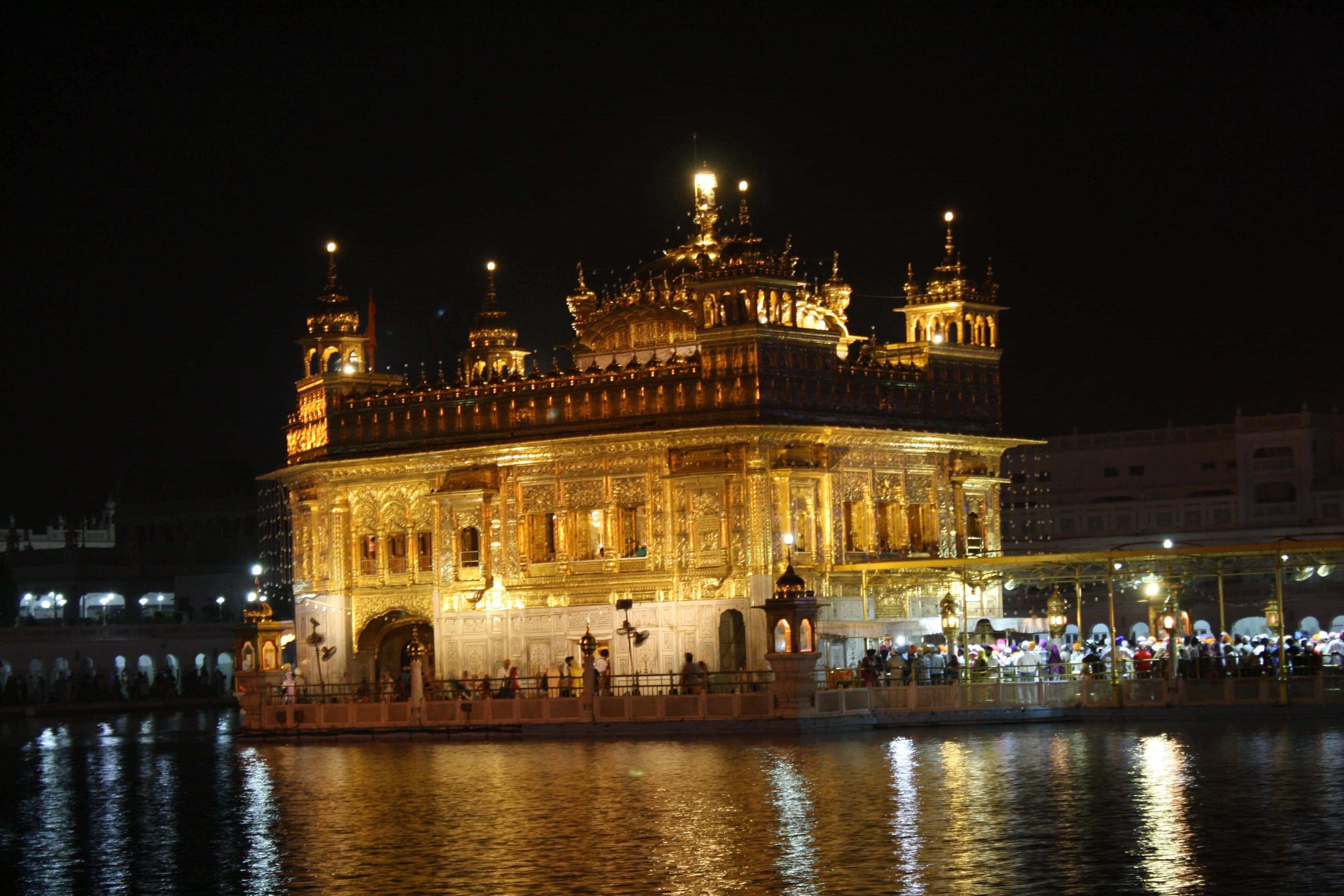 Golden Temple 桌布 壁紙 — 金色寺廟在夜晚被照亮，倒映在平靜的水面上，遊客聚集在其神聖的池塘周圍。