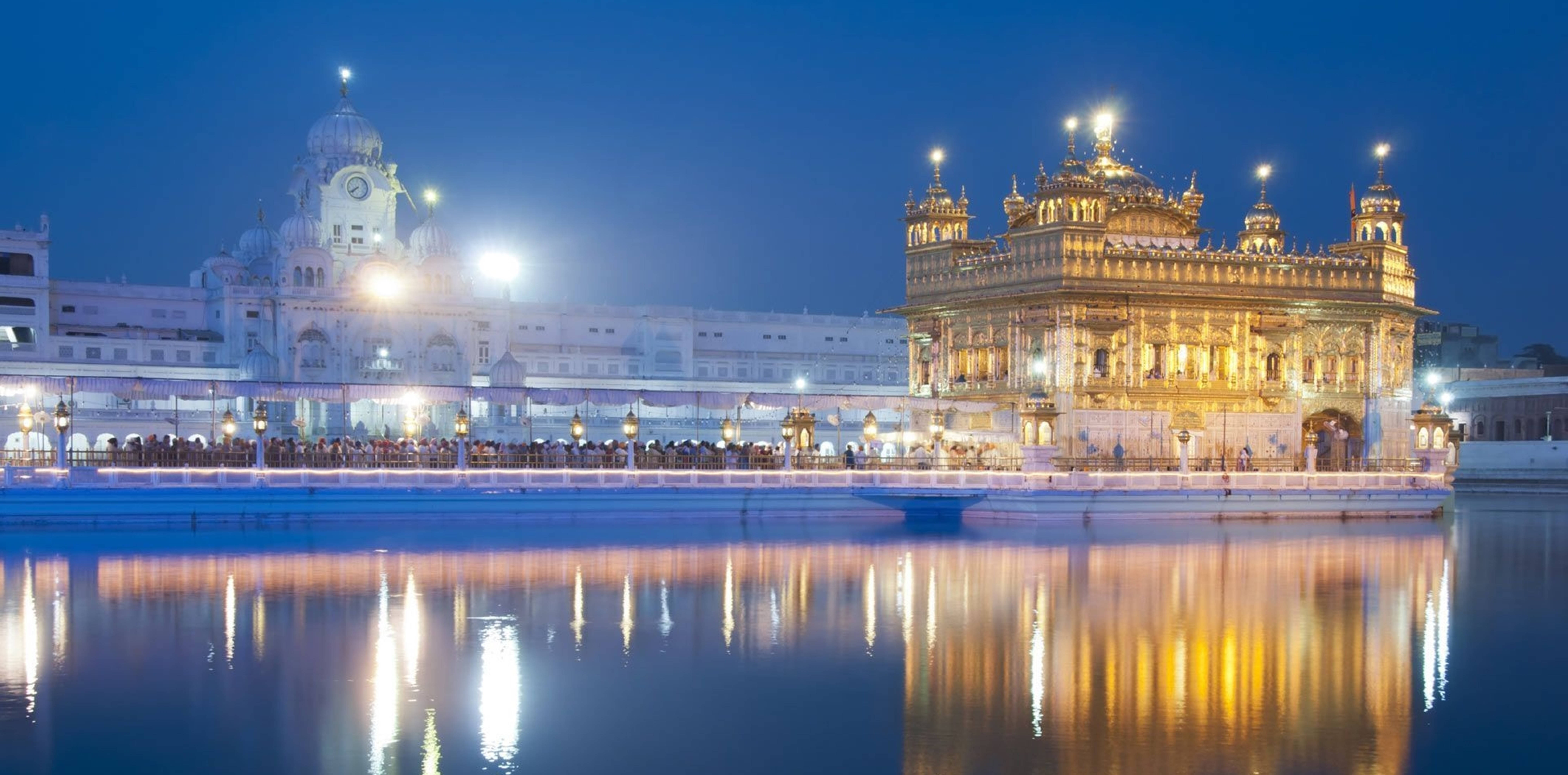 India 桌布 壁紙 — 金色寺廟在夜晚被照亮，發光的燈光倒映在平靜的水面上，周圍人群湧動。