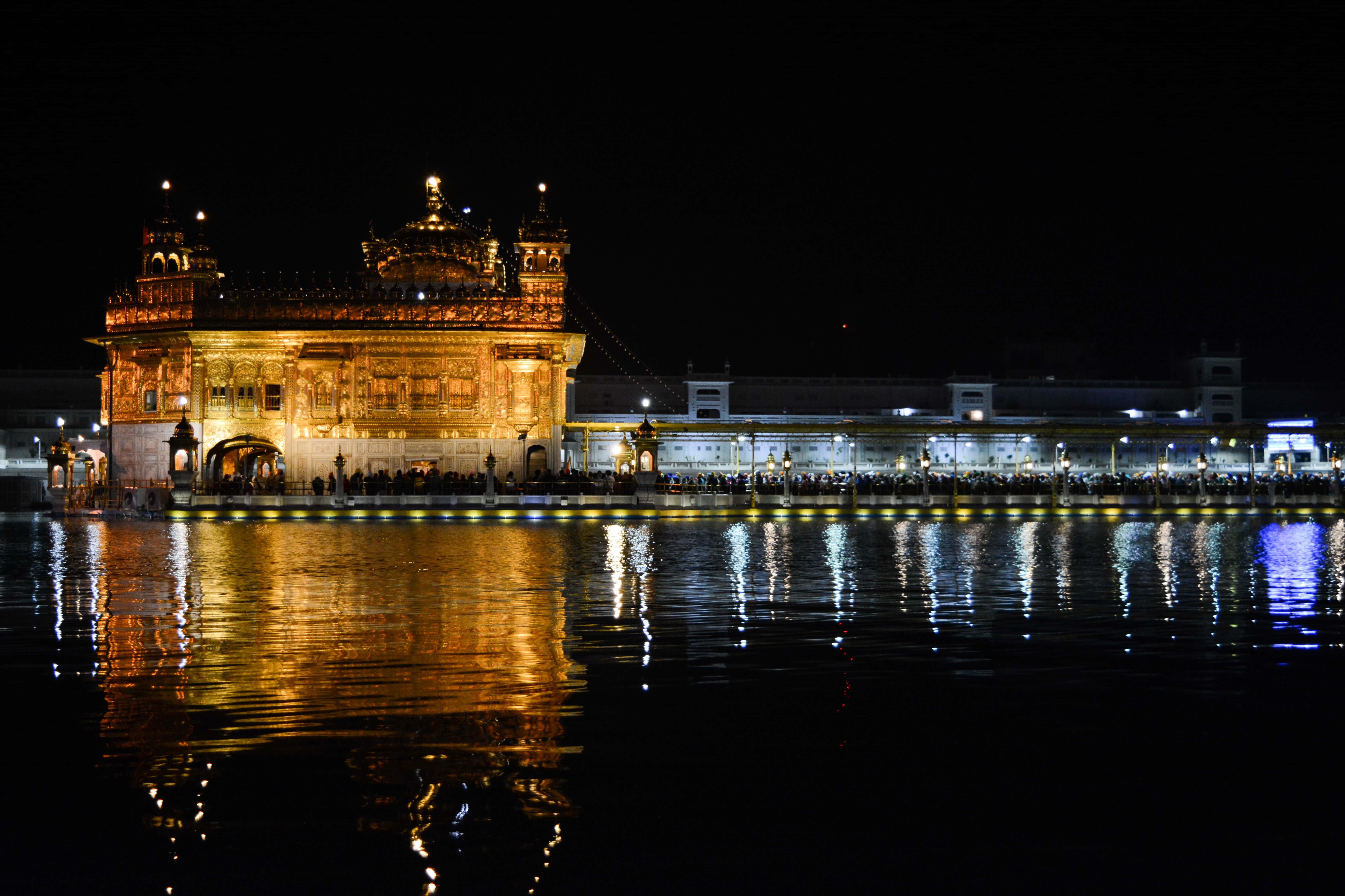 Golden Temple 桌布 壁紙 — 金色寺廟在夜間被溫暖的燈光照亮，倒映在平靜的水面上，周圍有遊客。