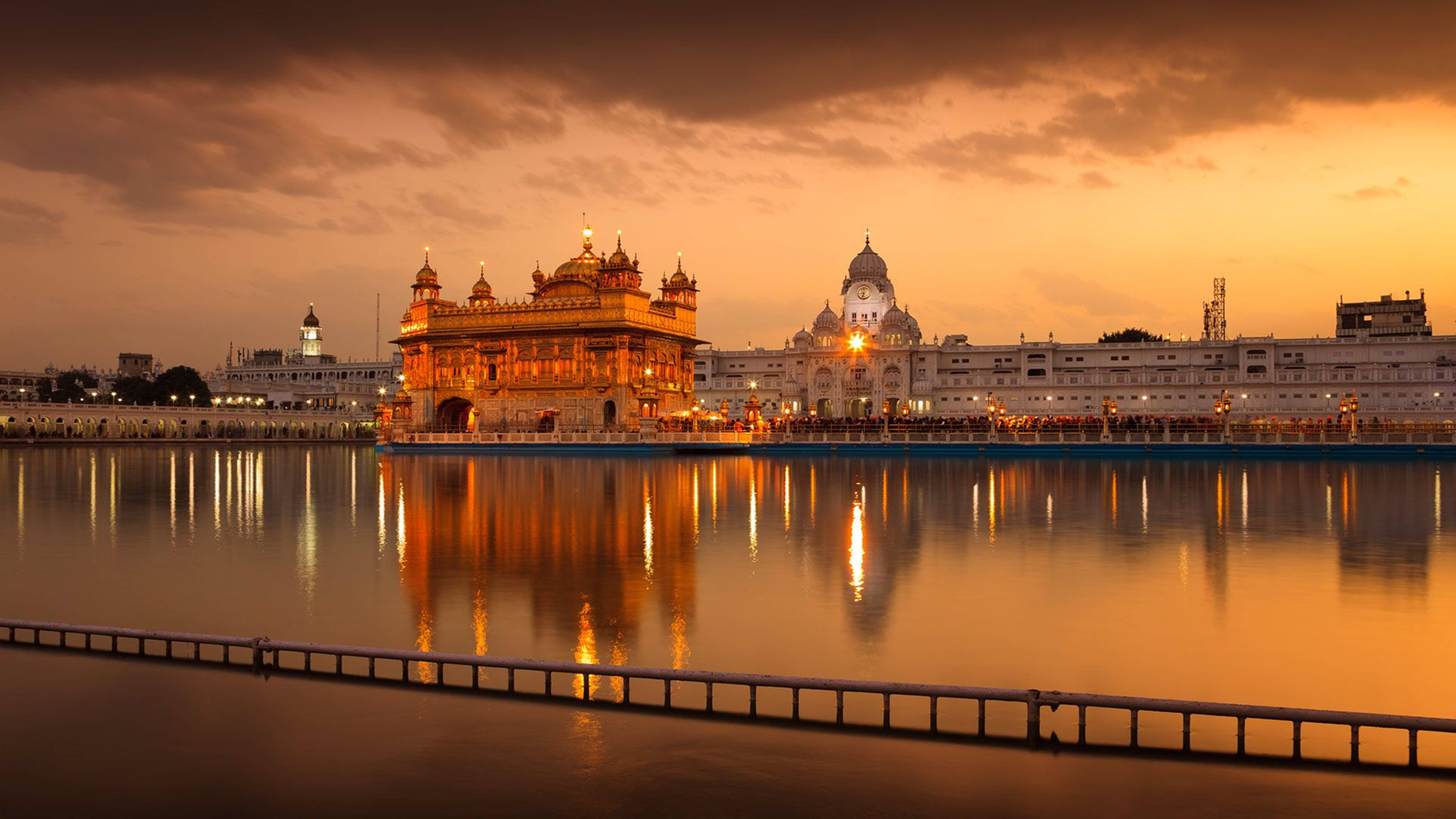 India 桌布 壁紙 — 金色寺廟在日落時溫暖地發光，其倒影在橙色雲朵下的平靜水面上閃爍。