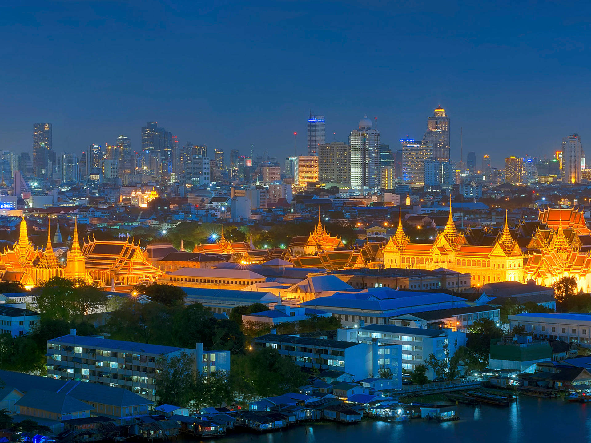 Bangkok 桌布 壁紙 — 金色寺廟尖頂在深藍色暮色天空下溫暖地發光，映襯著充滿活力的泰國城市景觀，有發光的摩天大樓和水面倒影。