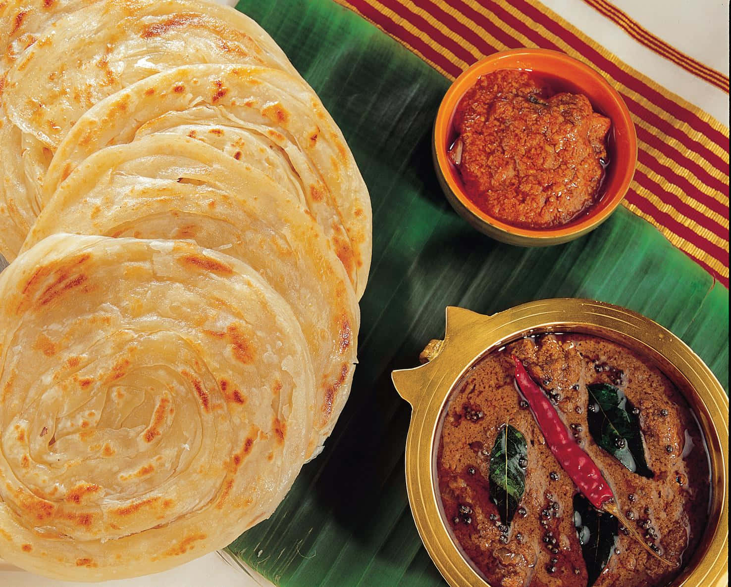 Indian 桌布 壁紙 — 金色層疊的印度煎餅堆疊在香蕉葉上，旁邊是裝飾碗中的辛辣咖哩和酸辣醬。