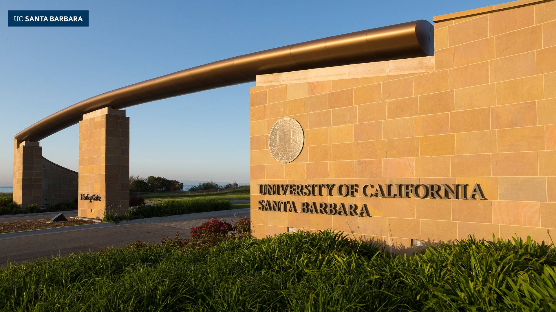 Architecture 桌布 壁紙 — 金色拱門和石牆上刻有「University of California Santa Barbara」，下方有綠色灌木，背景是清澈的藍天。
